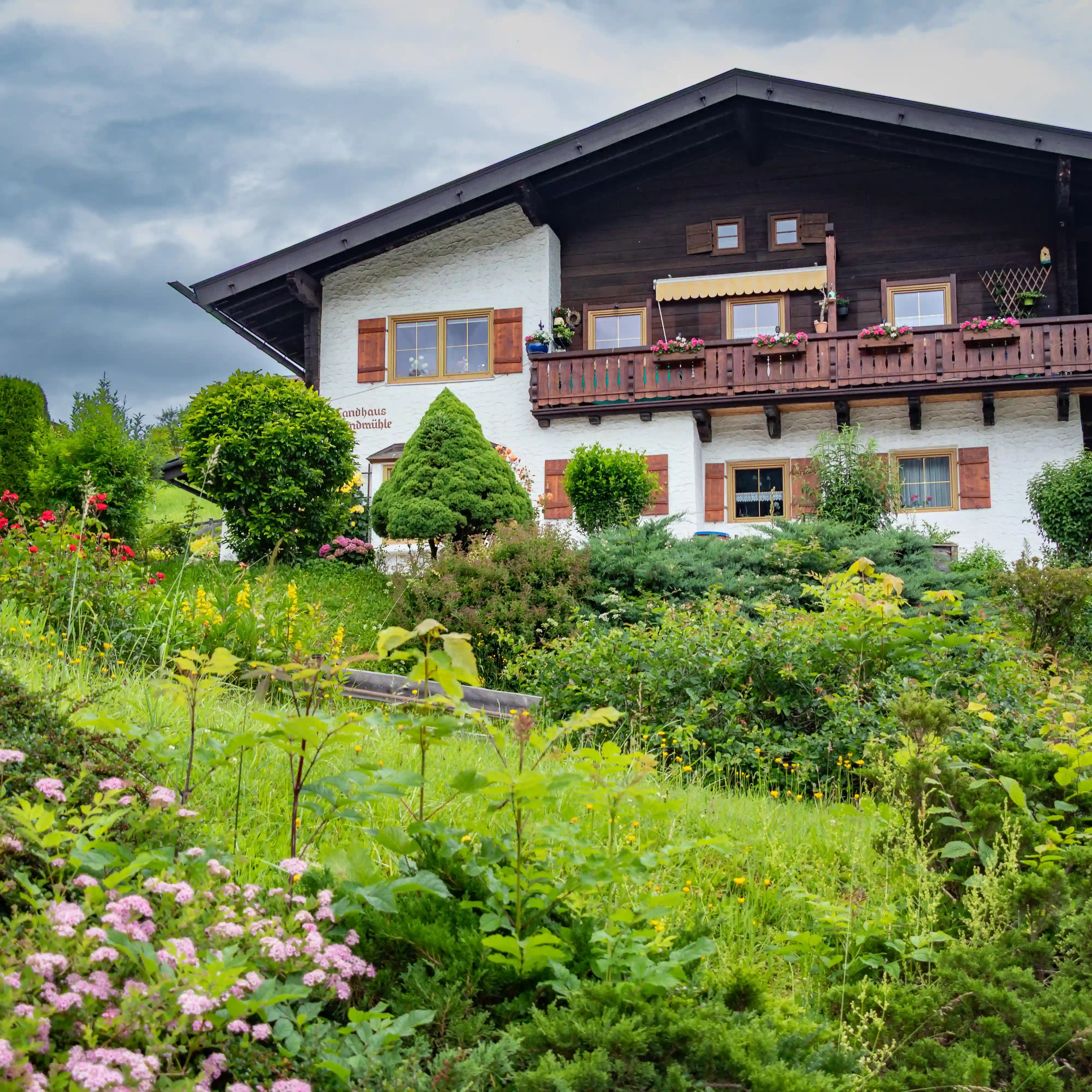 A traditional Alpine guesthouse with white walls, dark wood balconies, flower boxes, and a lush garden in the foreground.