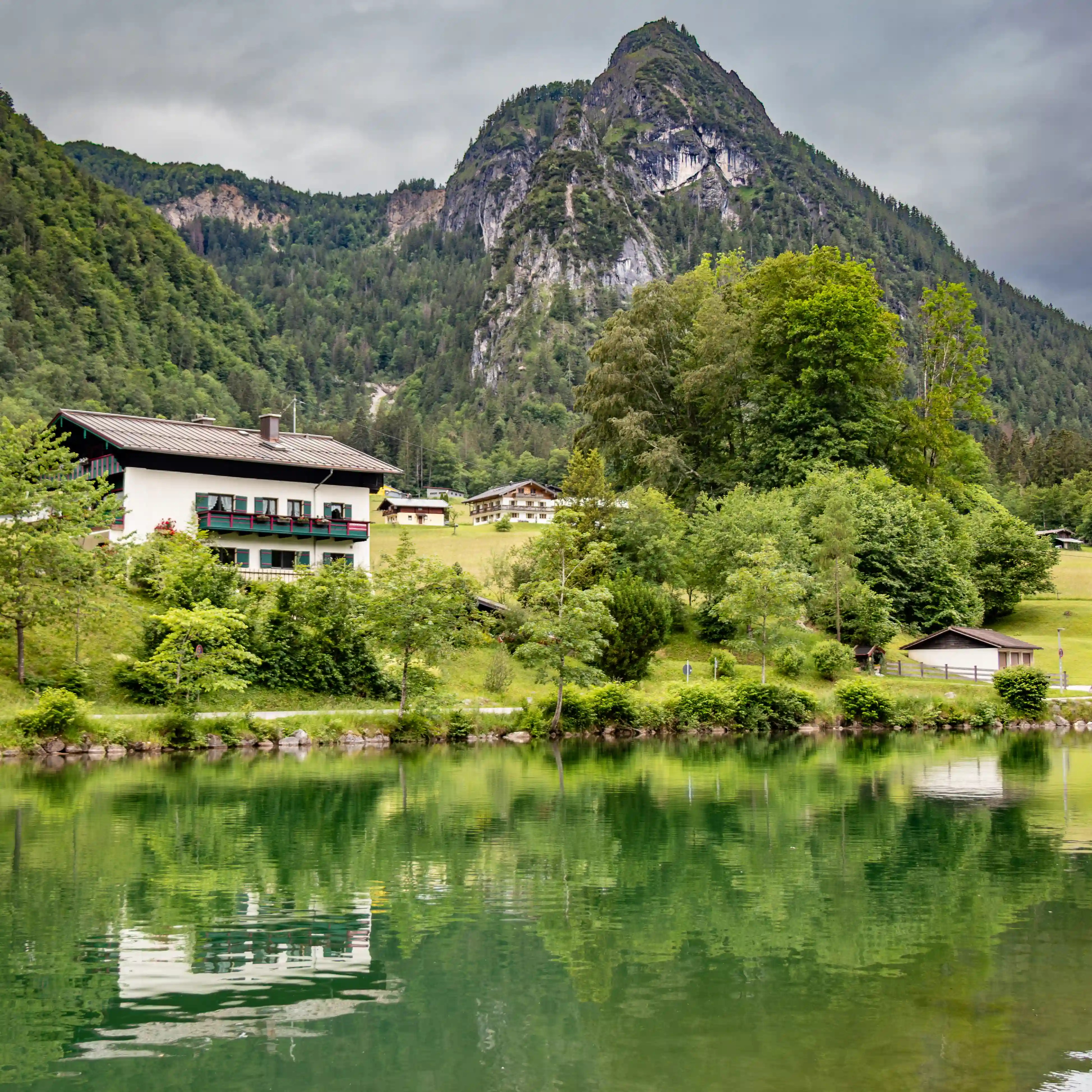 Green hills dotted with Alpine houses sit beneath a steep, rocky mountain rising sharply into a cloudy sky.
