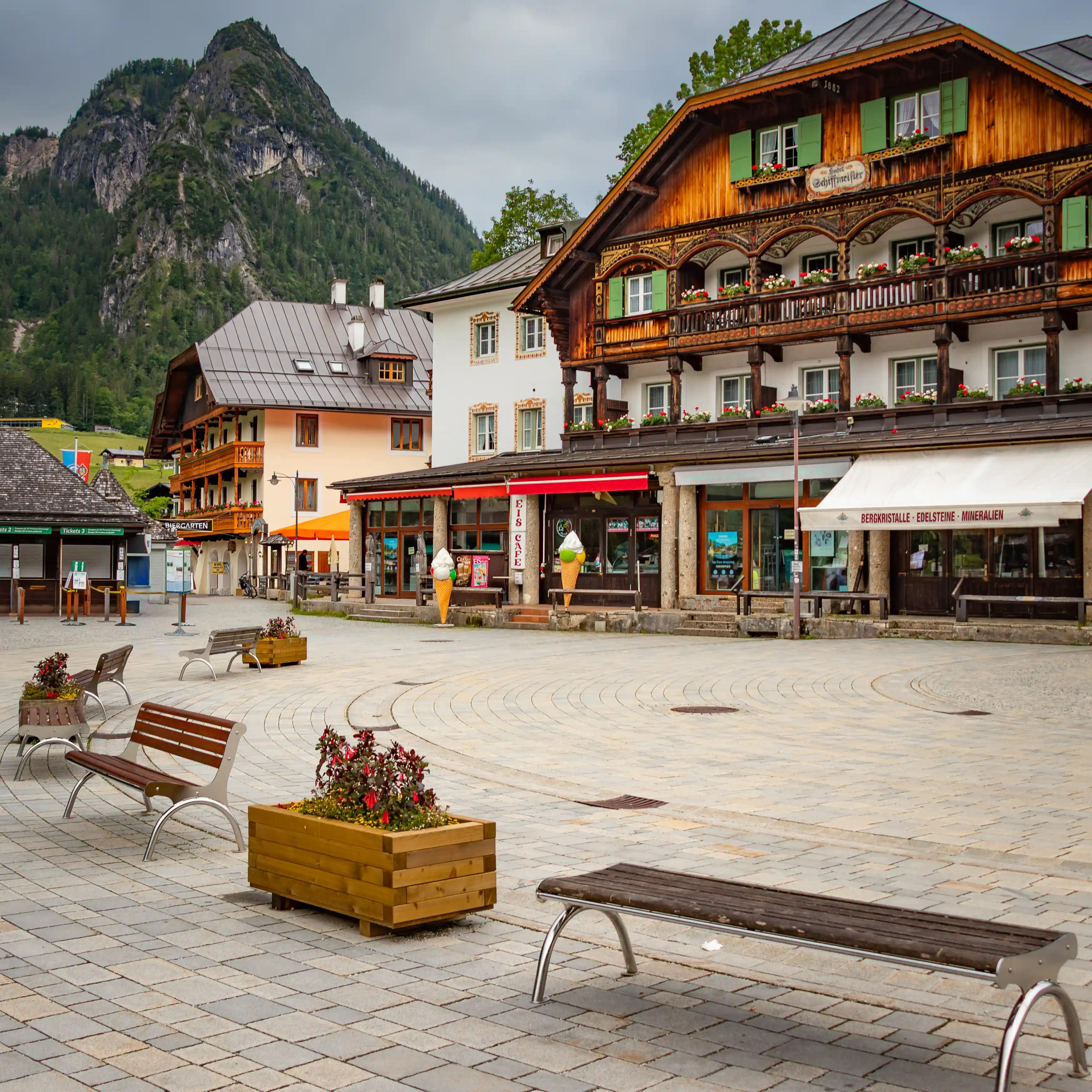 A wooden Alpine building with green shutters and flower boxes stands beside a small plaza with benches and shops.