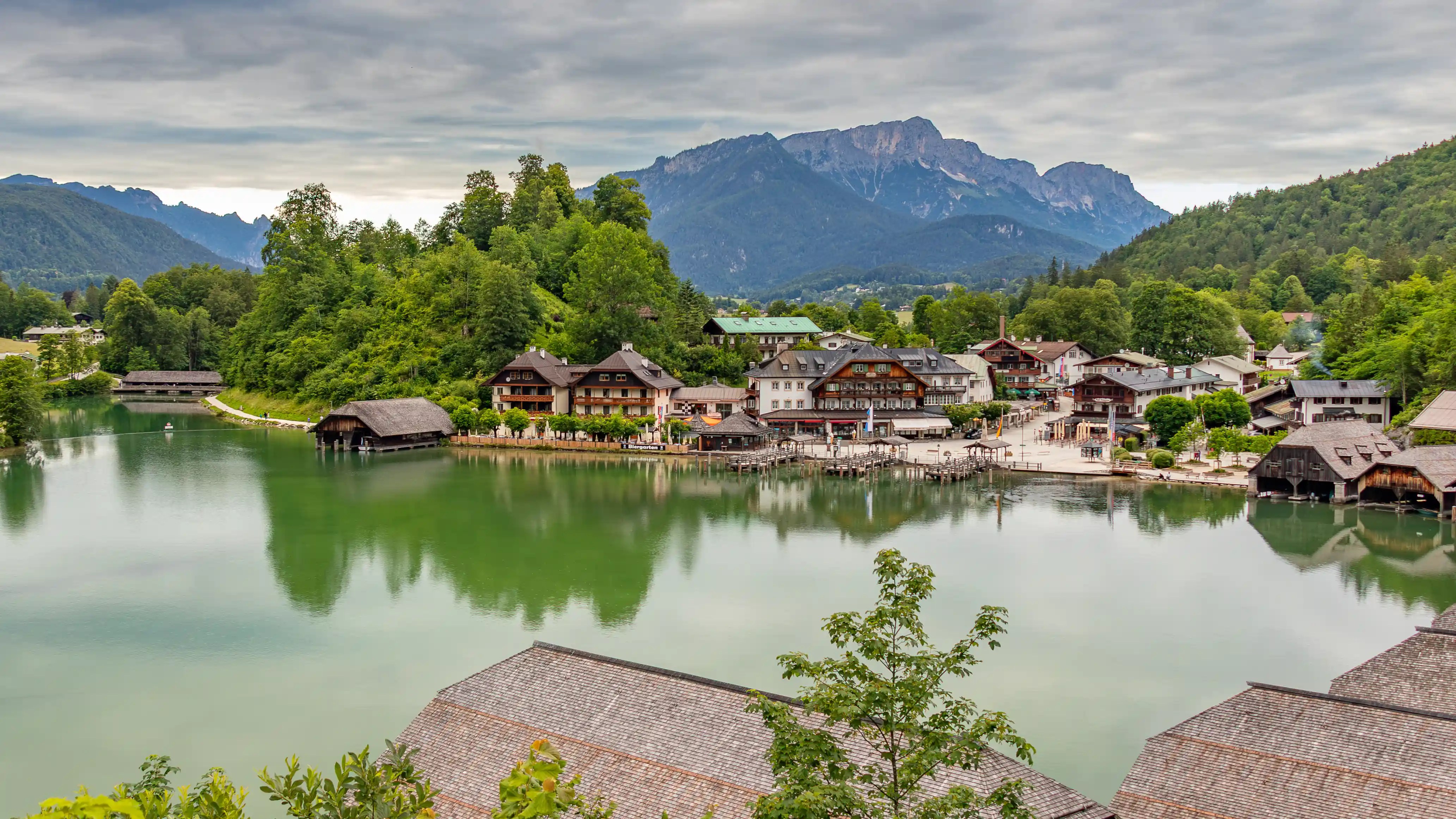 A wide view of emerald-green lake water reflects wooden boathouses and a small lakeside village surrounded by forested hills.