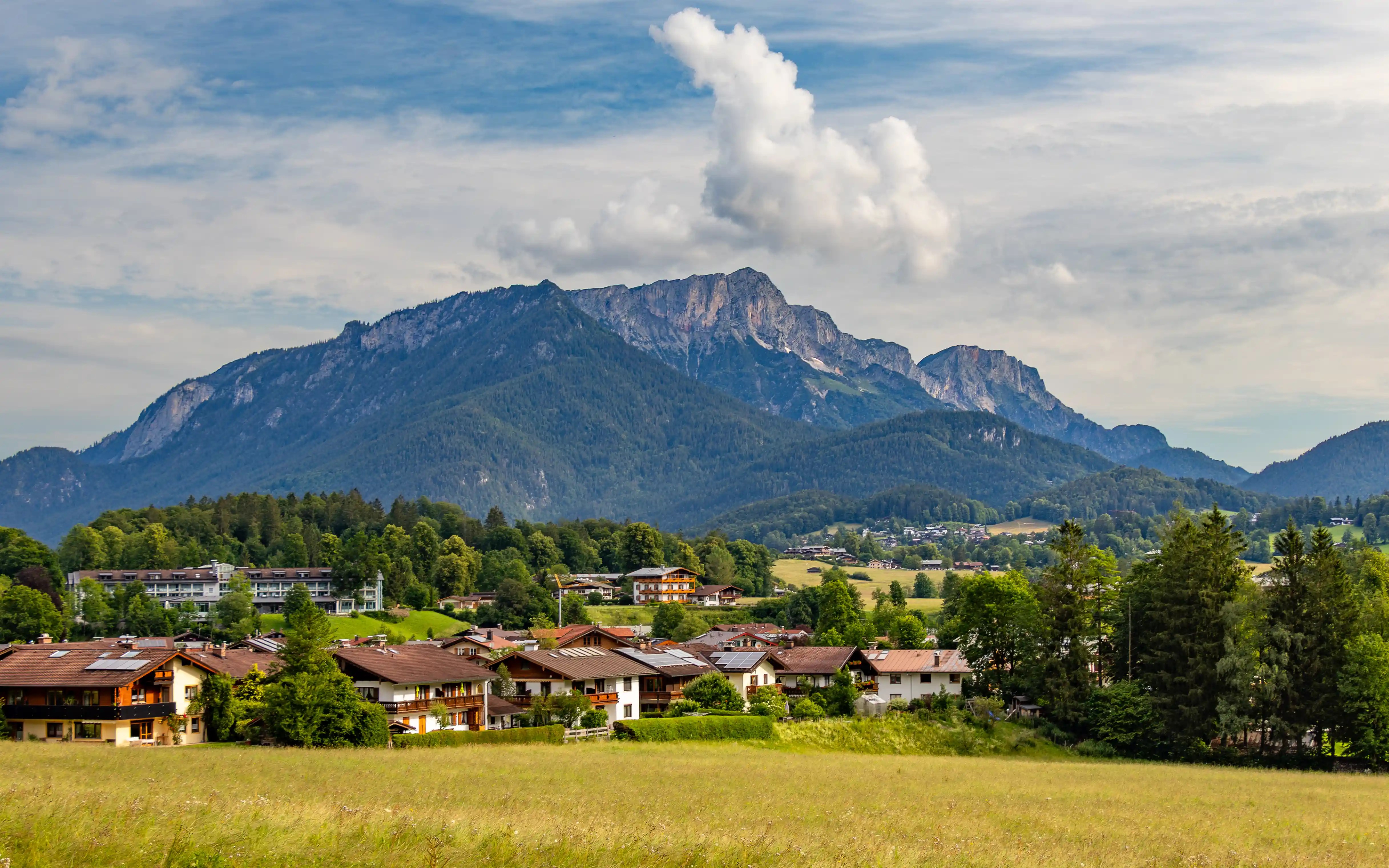 A broad mountain ridge rises behind a meadow and clustered Alpine houses under a partly cloudy sky.