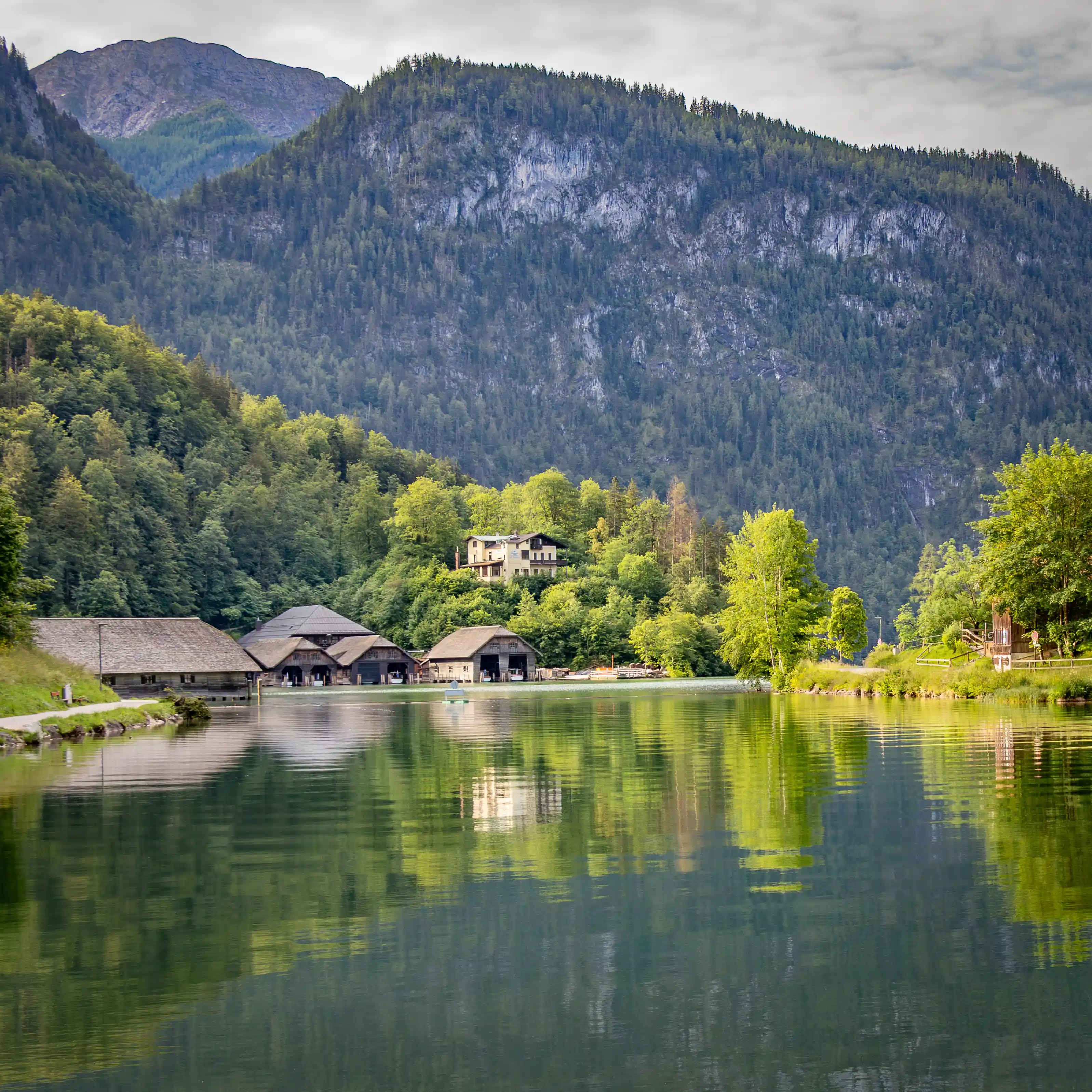 Wooden boathouses line the edge of Königssee, with green reflections rippling across the calm water and forested hills behind.