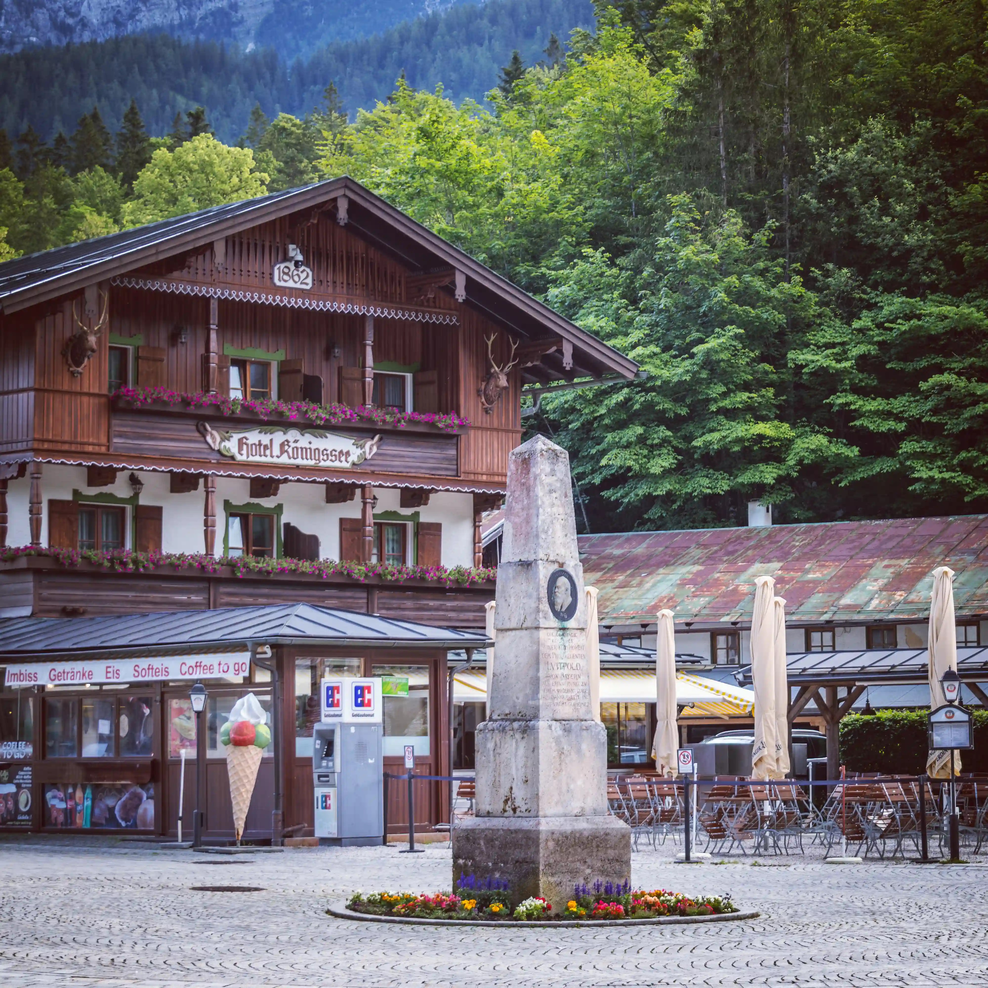 A wooden hotel building labeled “Hotel Königssee” stands beside a stone monument in a paved square.
