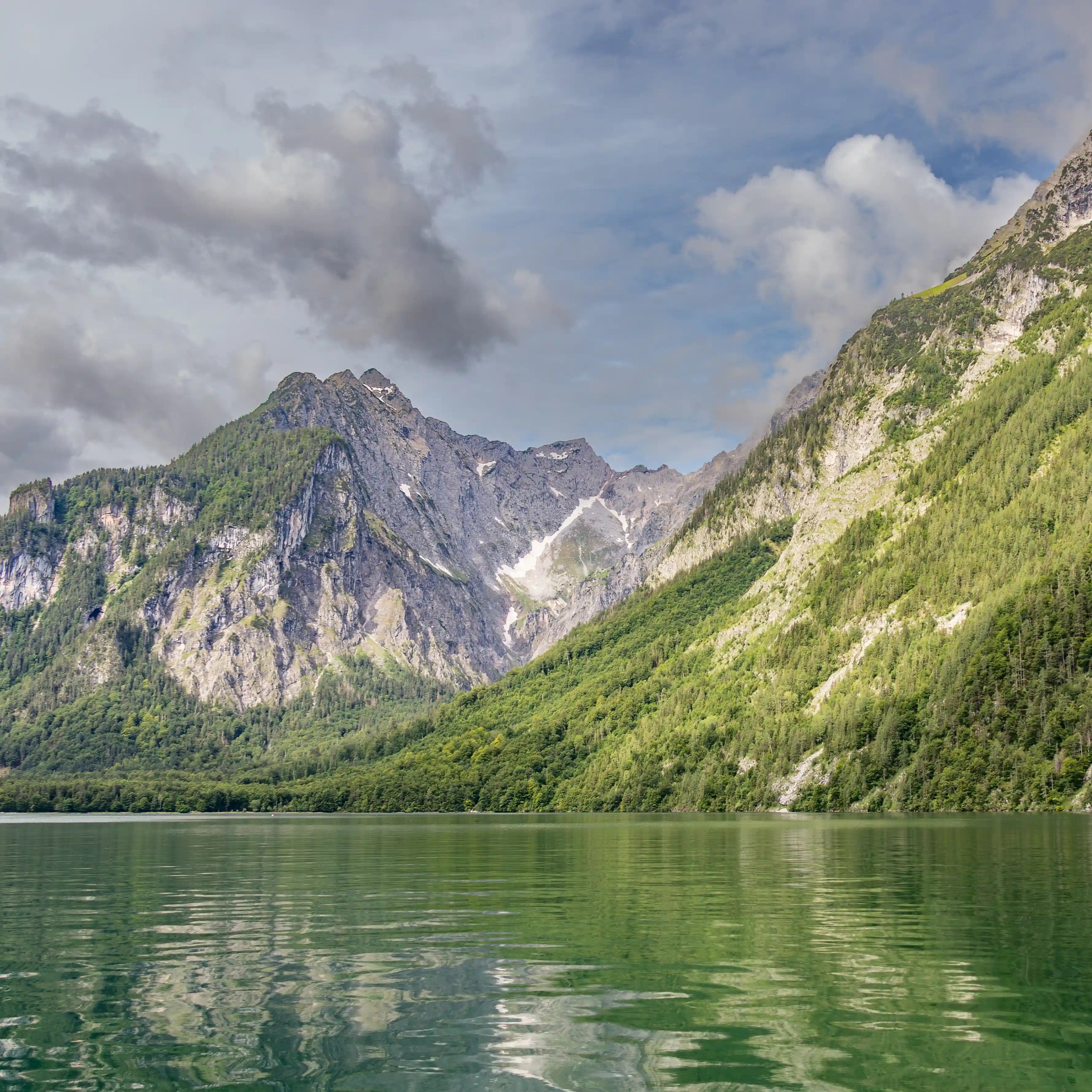 Steep rocky mountains covered in trees reflect clearly in the calm green water of Königssee.