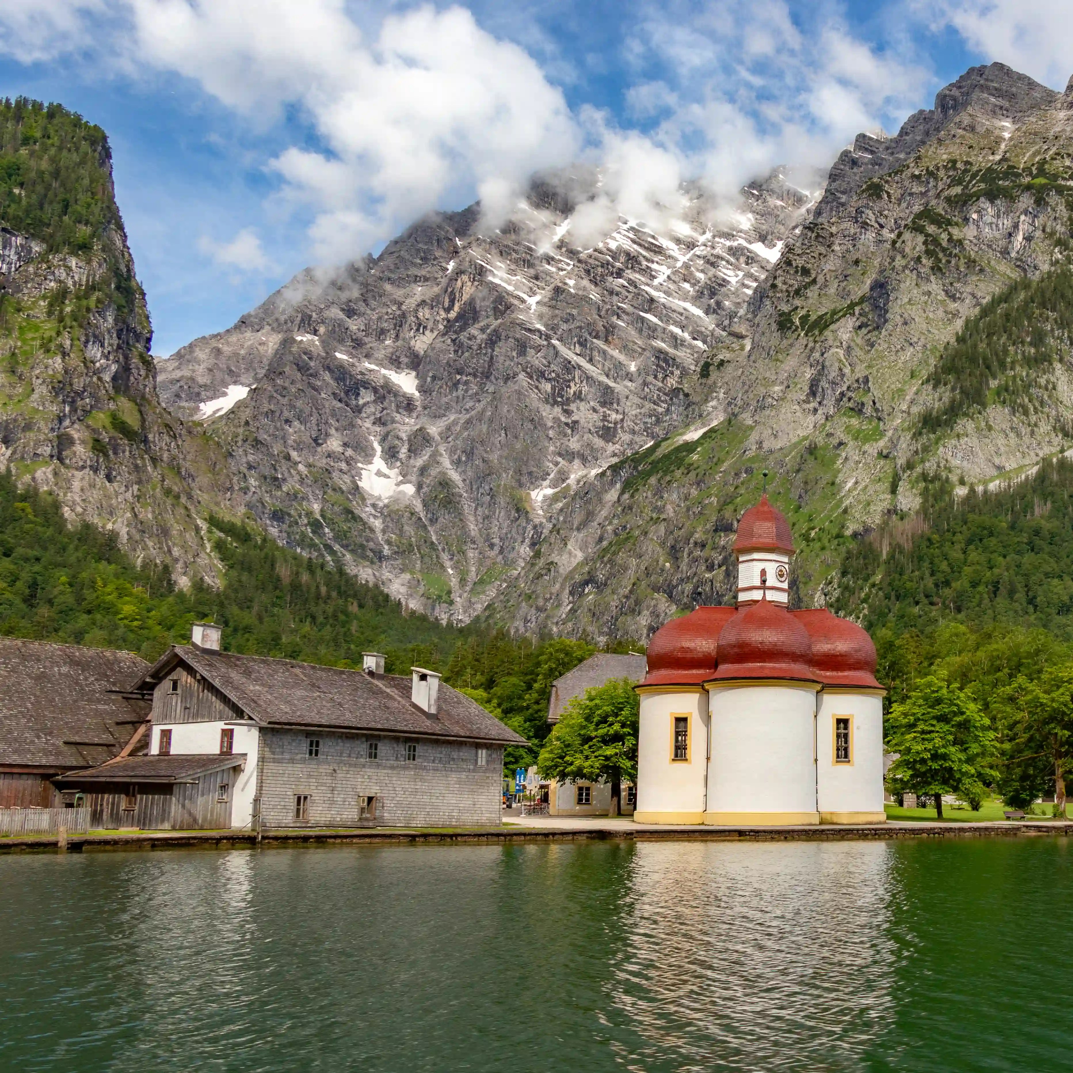 The Church of St. Bartholomä and nearby wooden buildings reflect in the green waters of Königssee beneath rugged mountains.