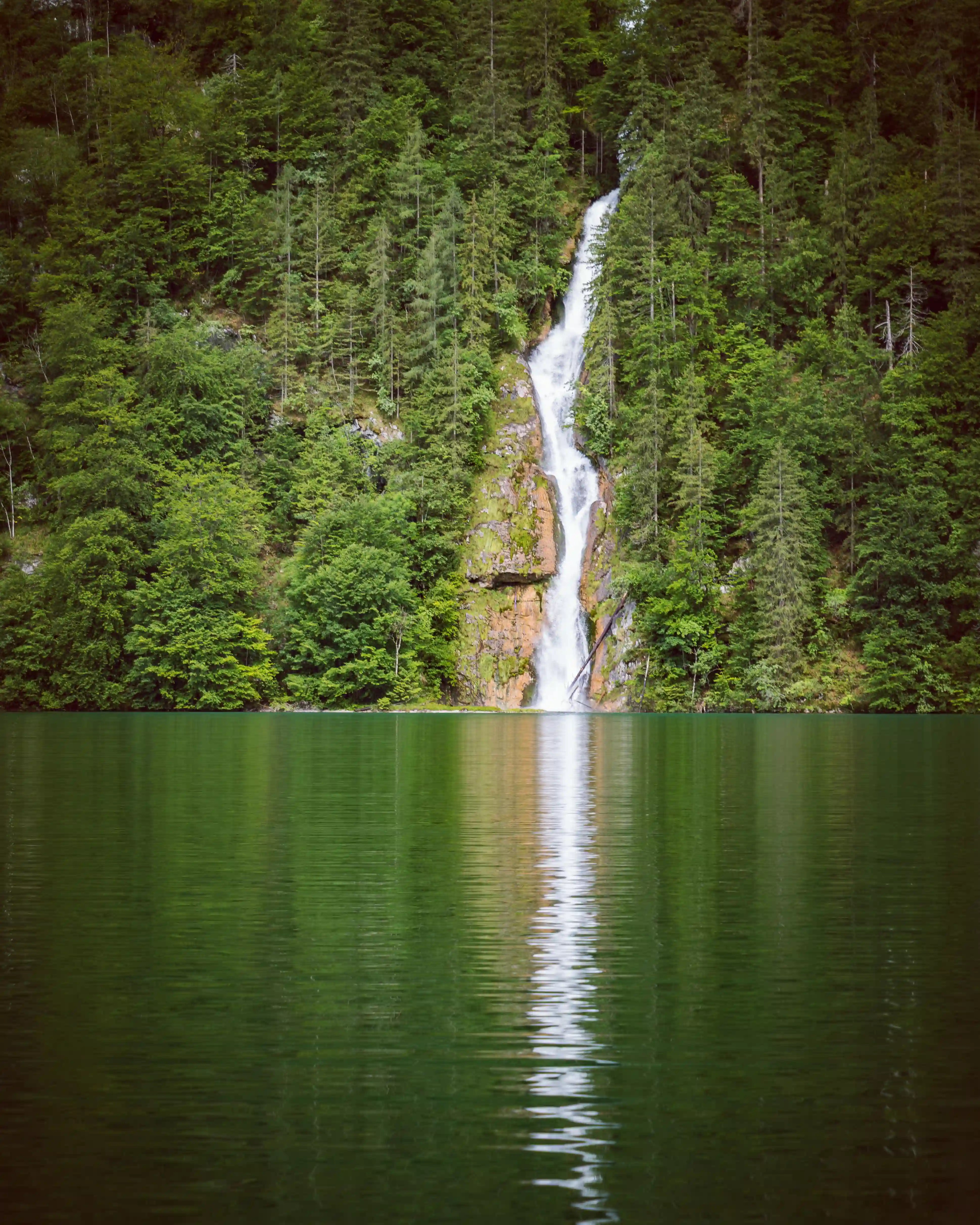 A narrow waterfall pours down a steep forested cliff and flows directly into the emerald-green waters of Königssee.