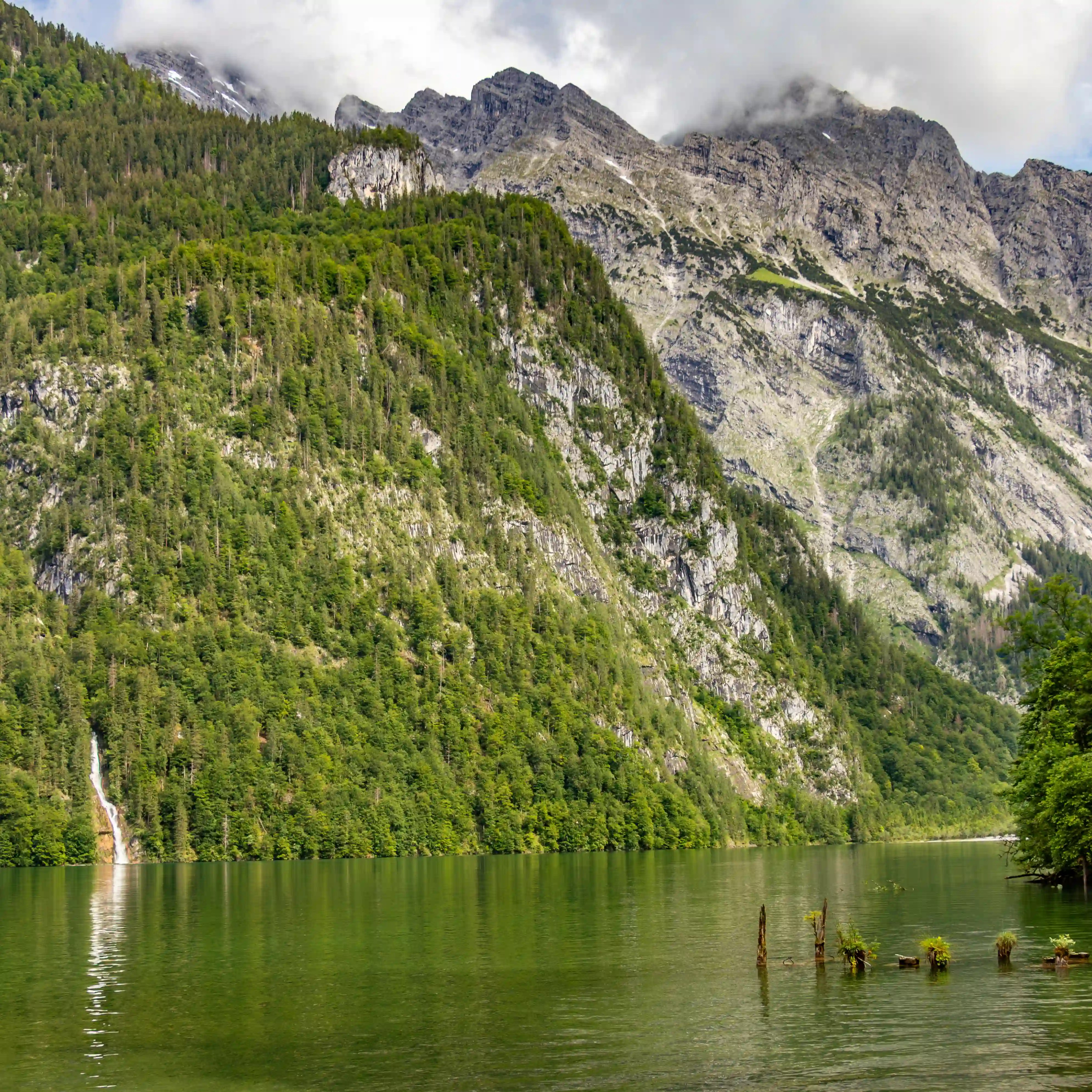 Calm green water stretches across Königssee with steep forested mountains and a thin waterfall visible in the distance.