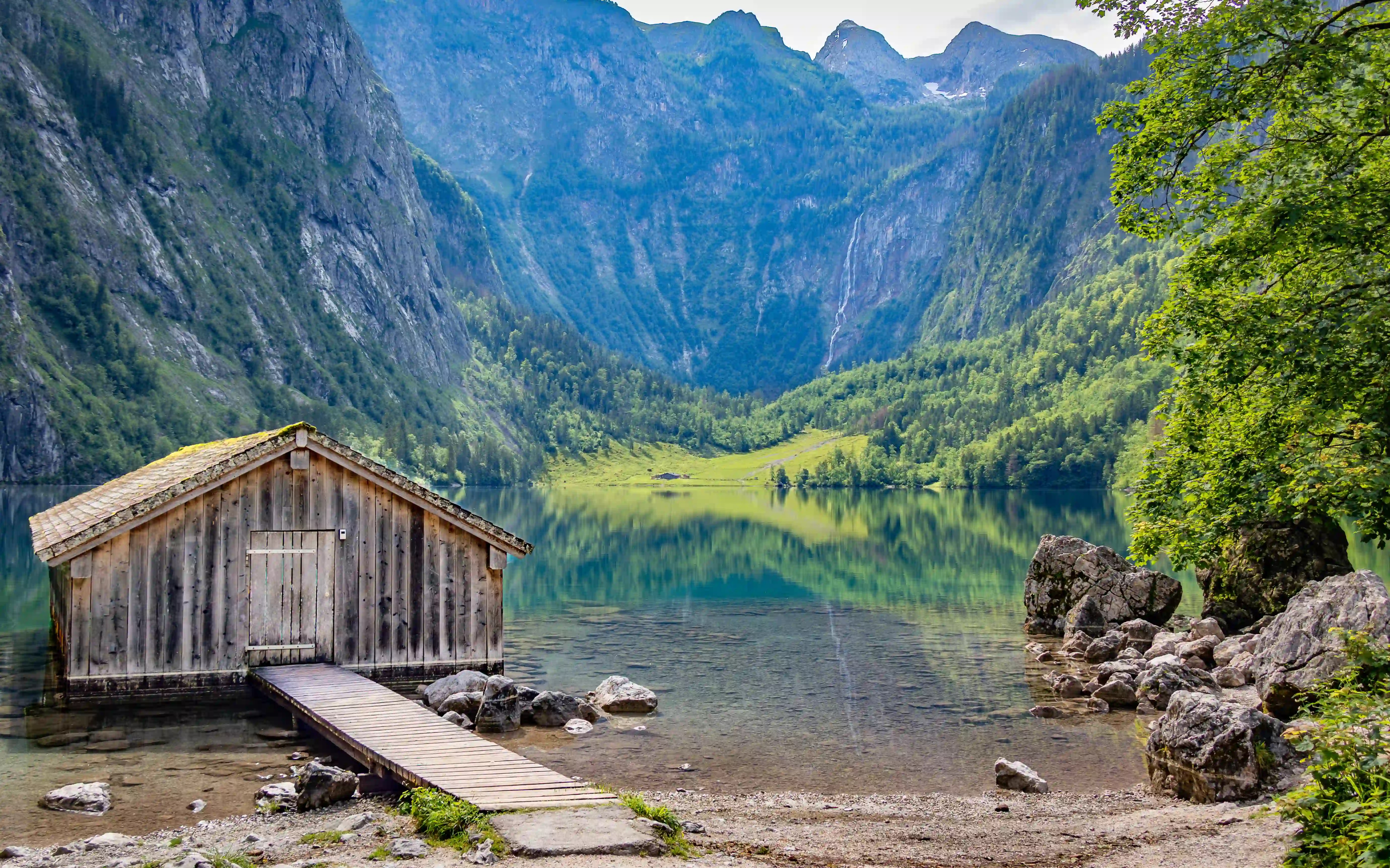 A small wooden boathouse sits at the water’s edge with a narrow dock extending into the clear lake, backed by steep forested mountains.