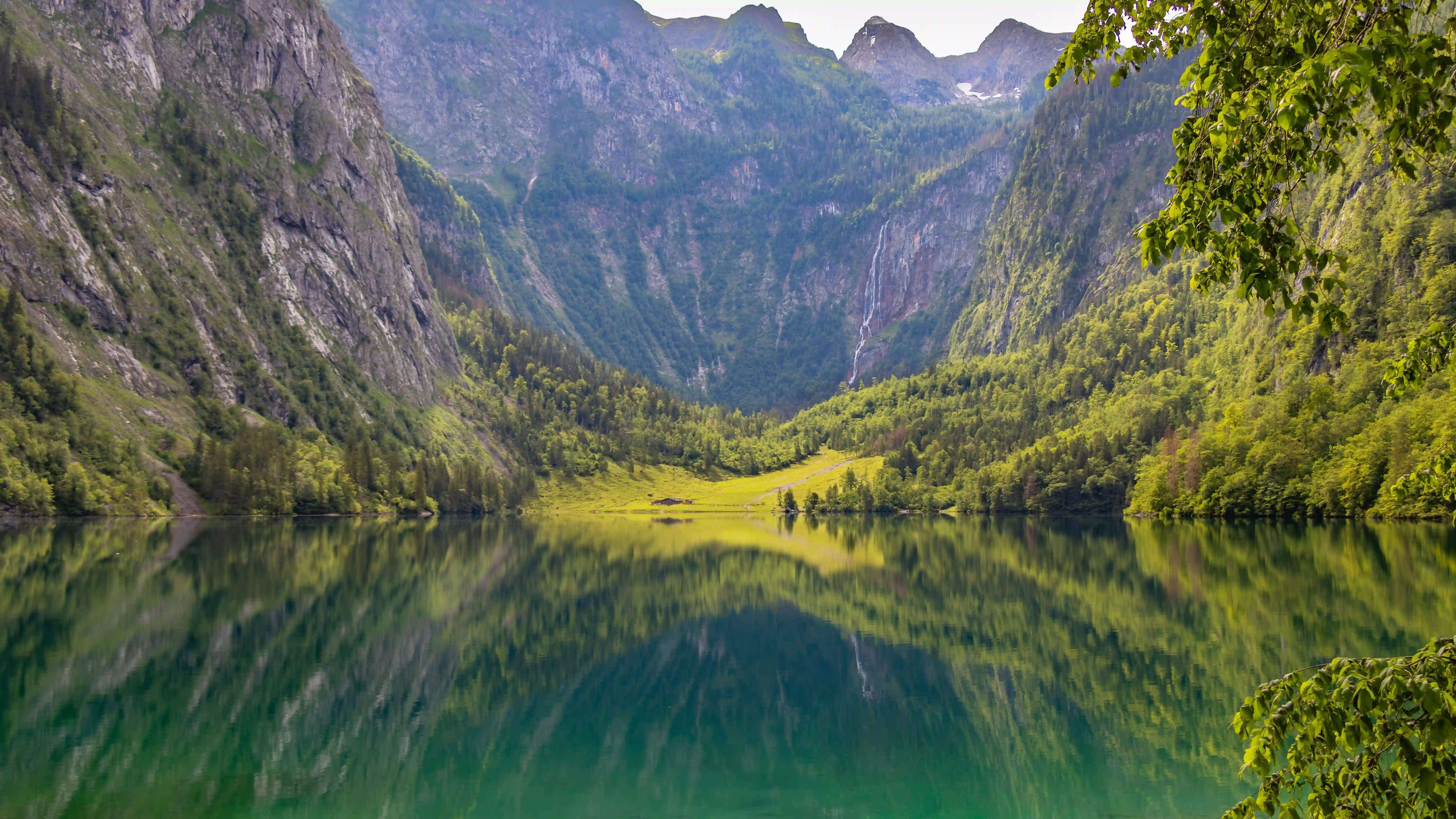 A wide view of Obersee shows green mountains and a bright meadow reflected in the still lake surface.