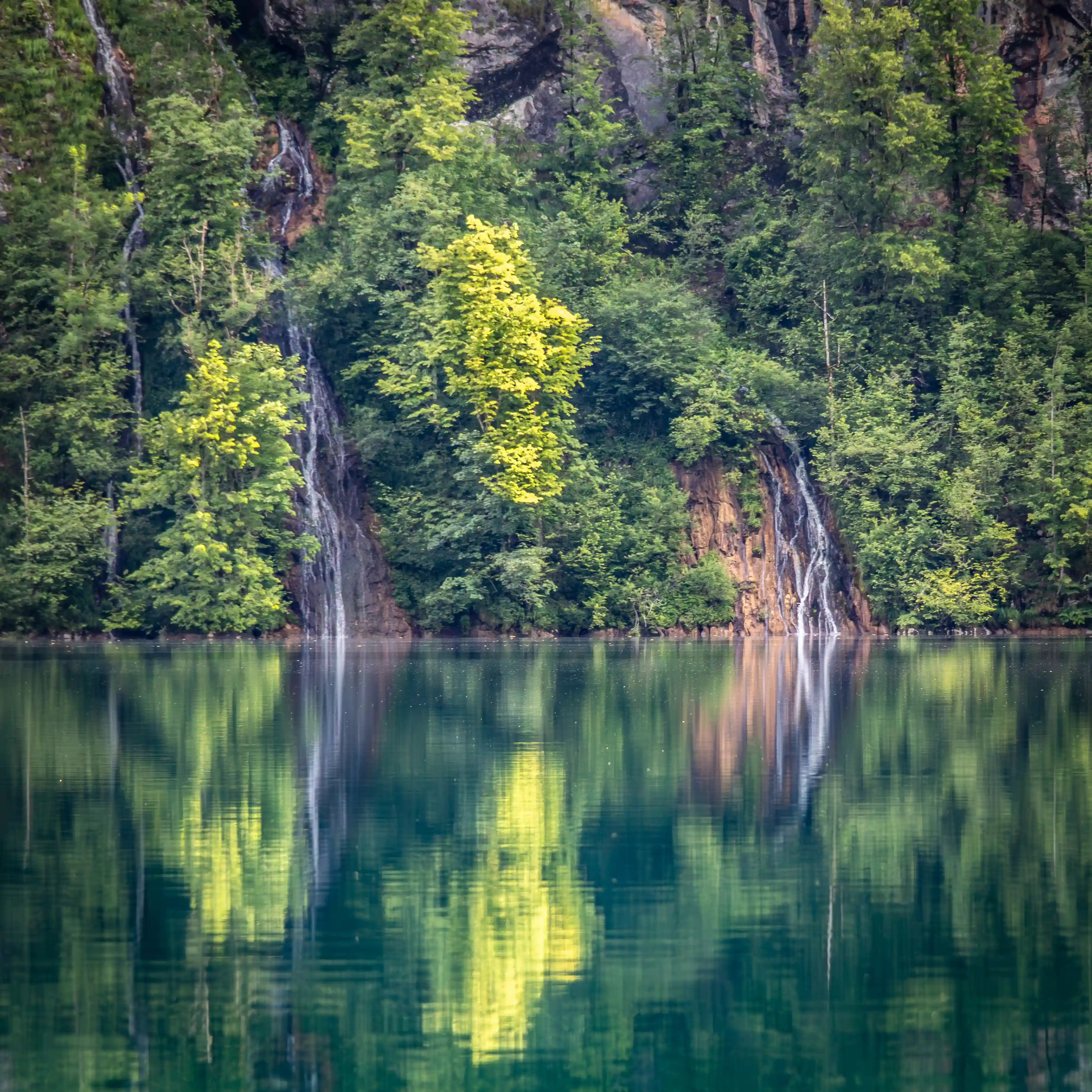 Two narrow waterfalls cascade down a forested rock face and reflect in the calm water below.