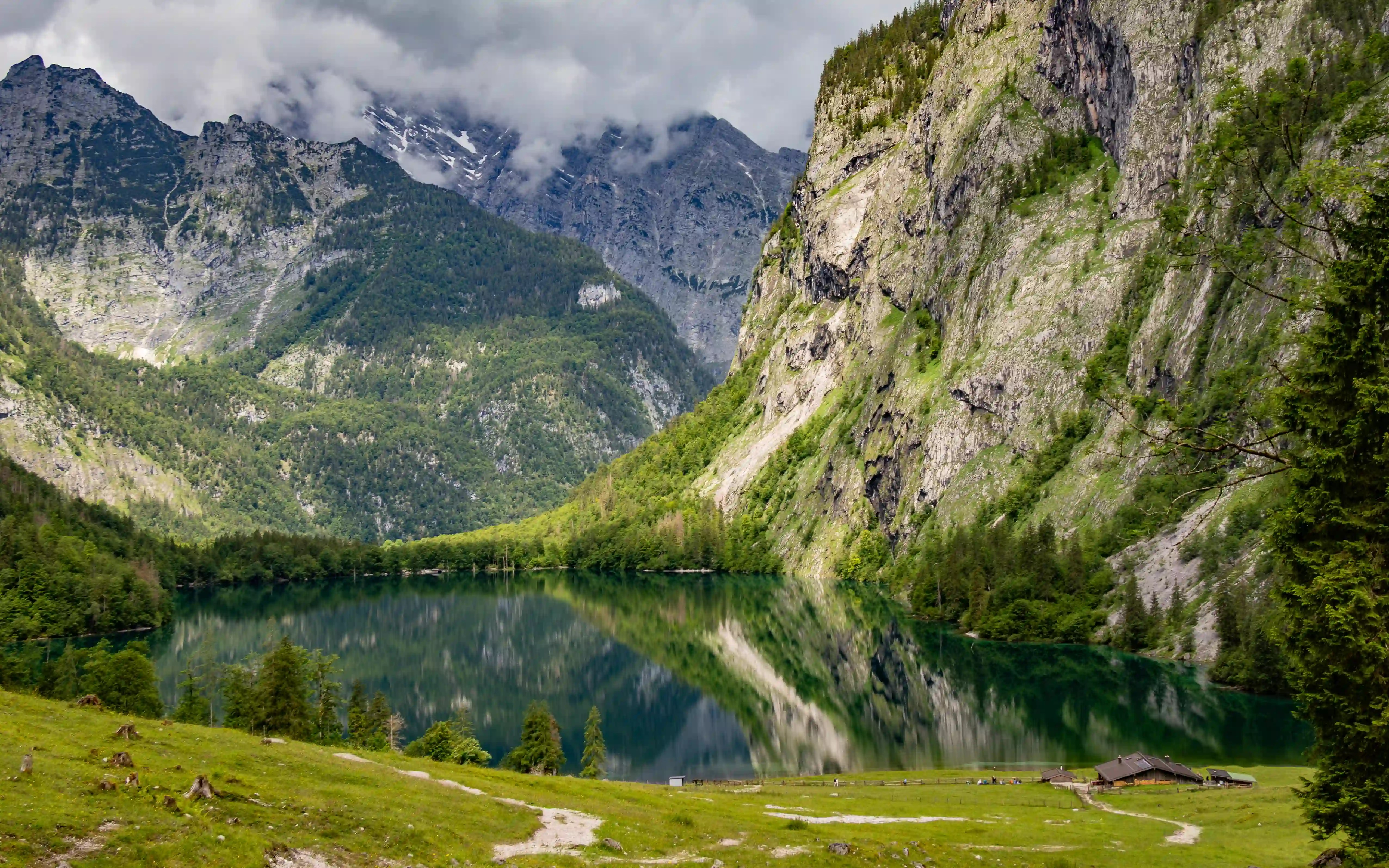 A calm alpine lake reflects steep forested cliffs and cloud-covered mountain peaks under an overcast sky.