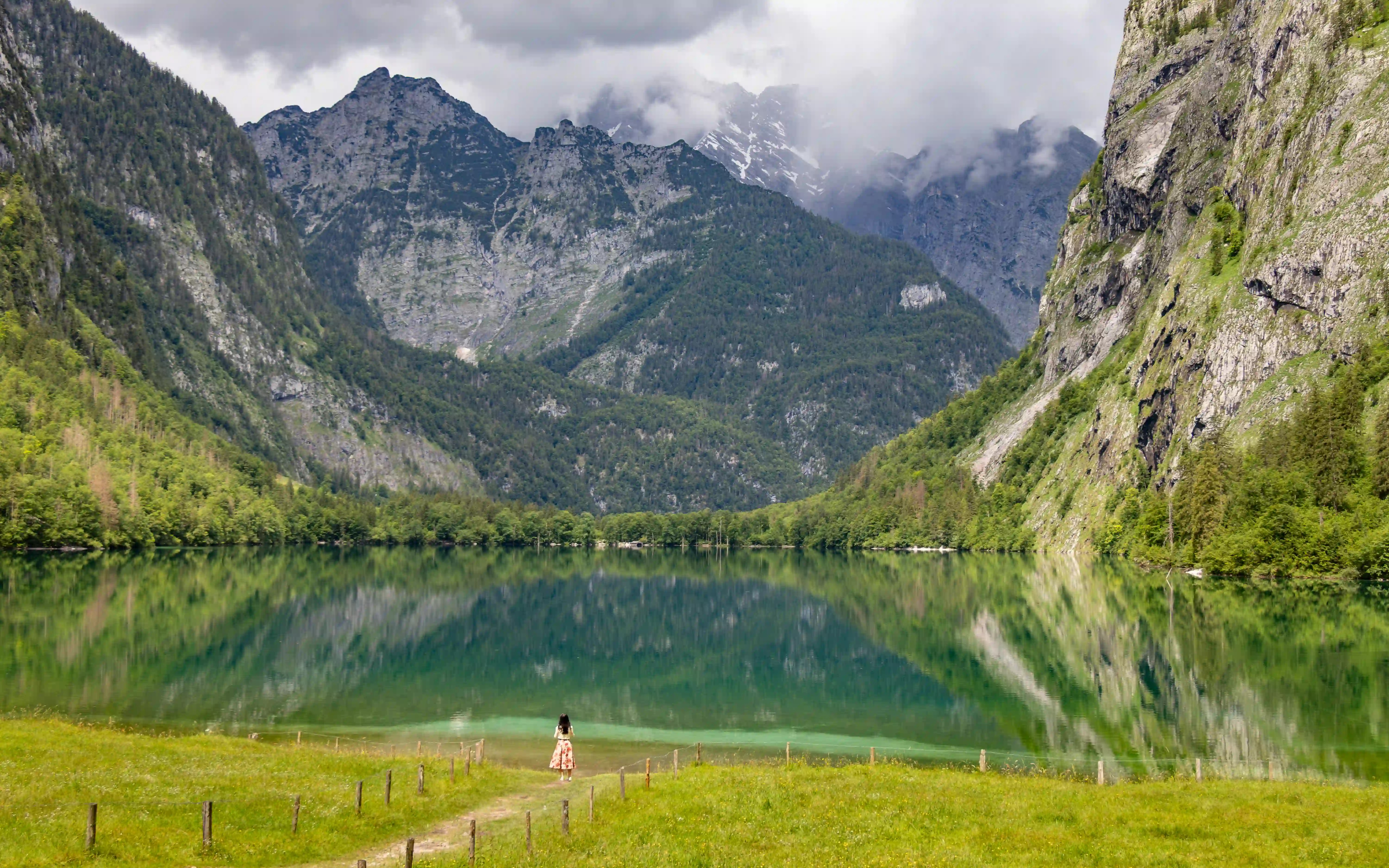 A narrow path leads through a grassy meadow toward the edge of Obersee, where a person stands facing the water and mountains beyond.