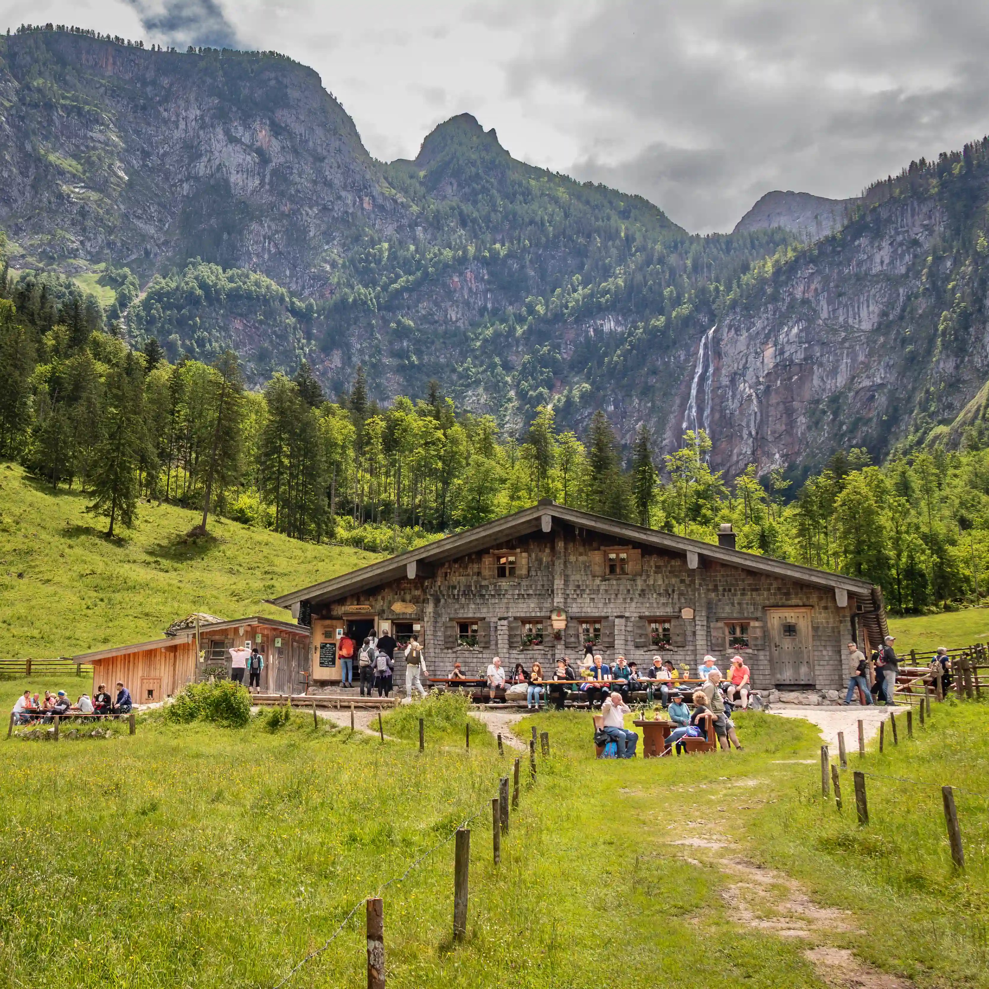 A wooden alpine hut sits in a green meadow with people gathered outside, while Röthbachfall cascades down a steep cliff in the background.