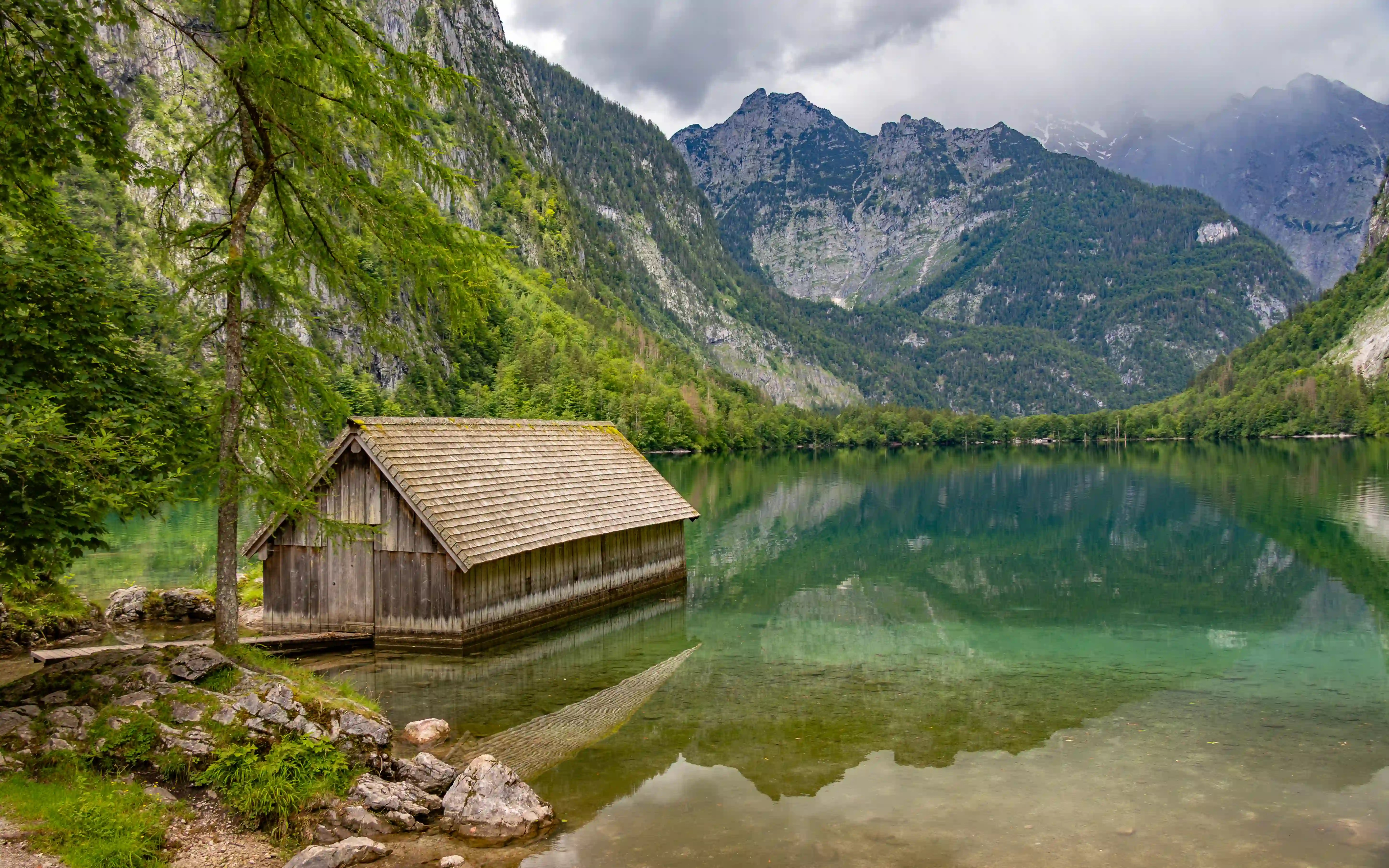 A small wooden boathouse rests at the edge of Obersee, reflected in the turquoise water with forested cliffs rising behind it.