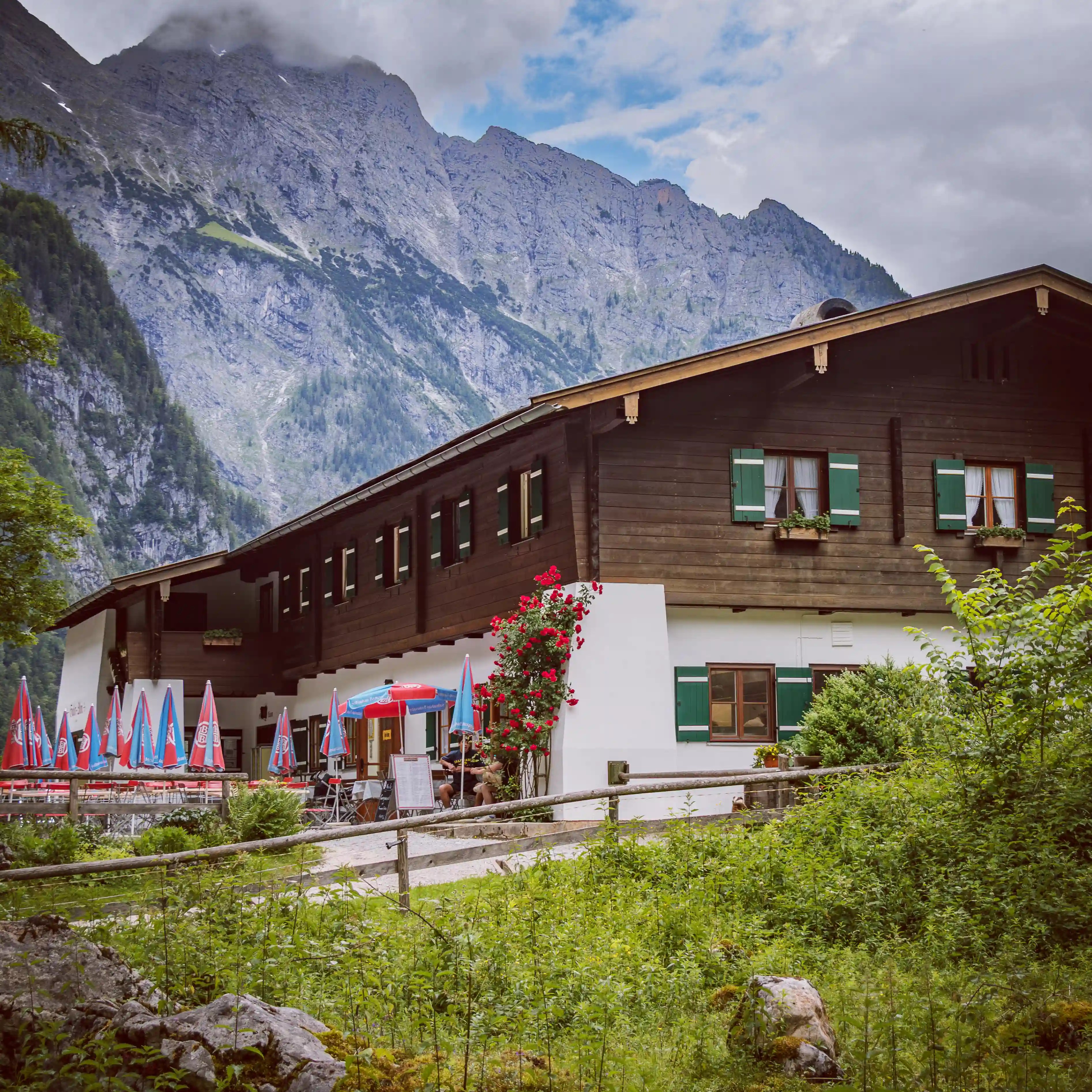 A traditional alpine restaurant with green shutters and outdoor seating sits beneath towering rocky mountains.