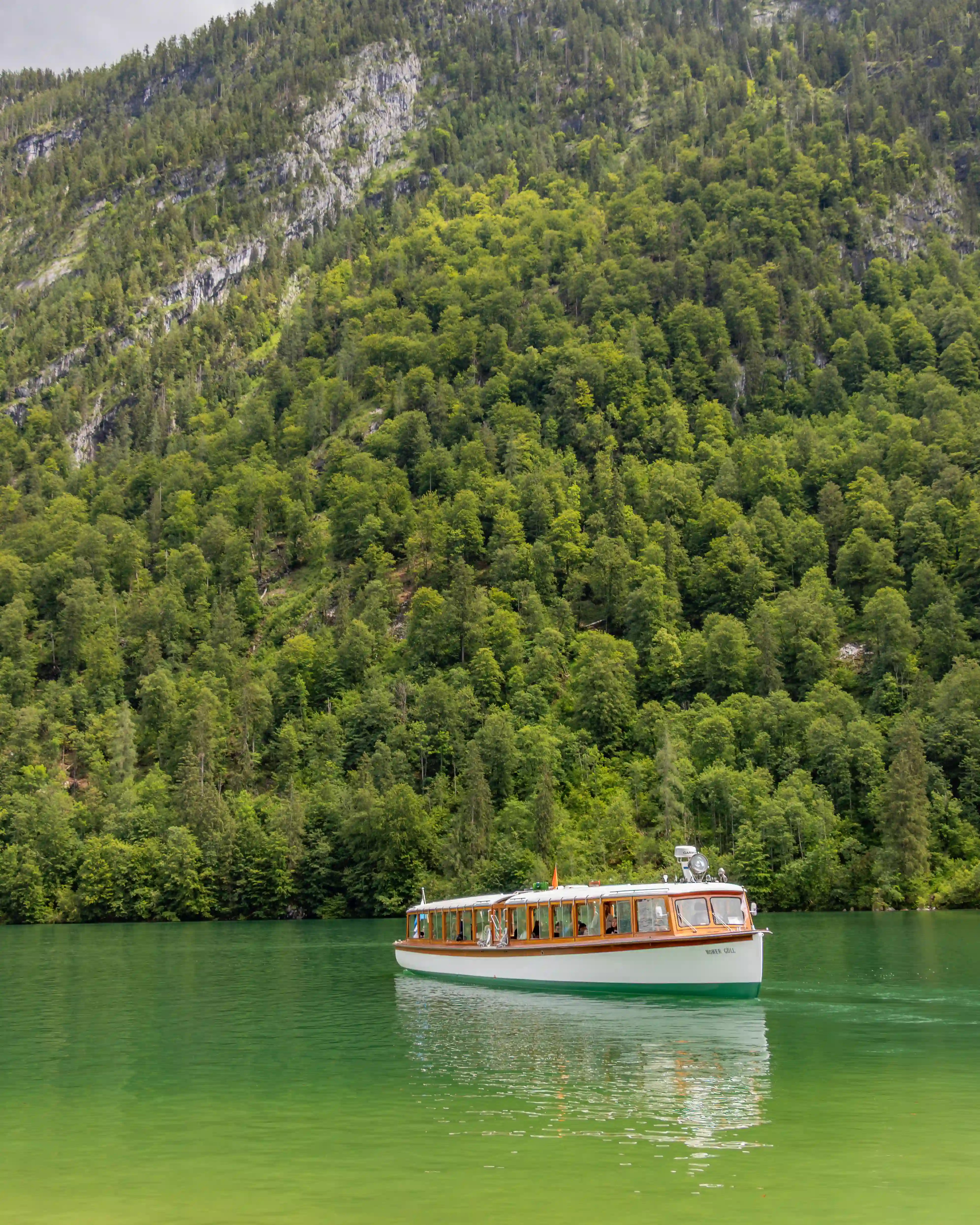 A traditional electric passenger boat glides across emerald-green water with a forested mountainside behind it.