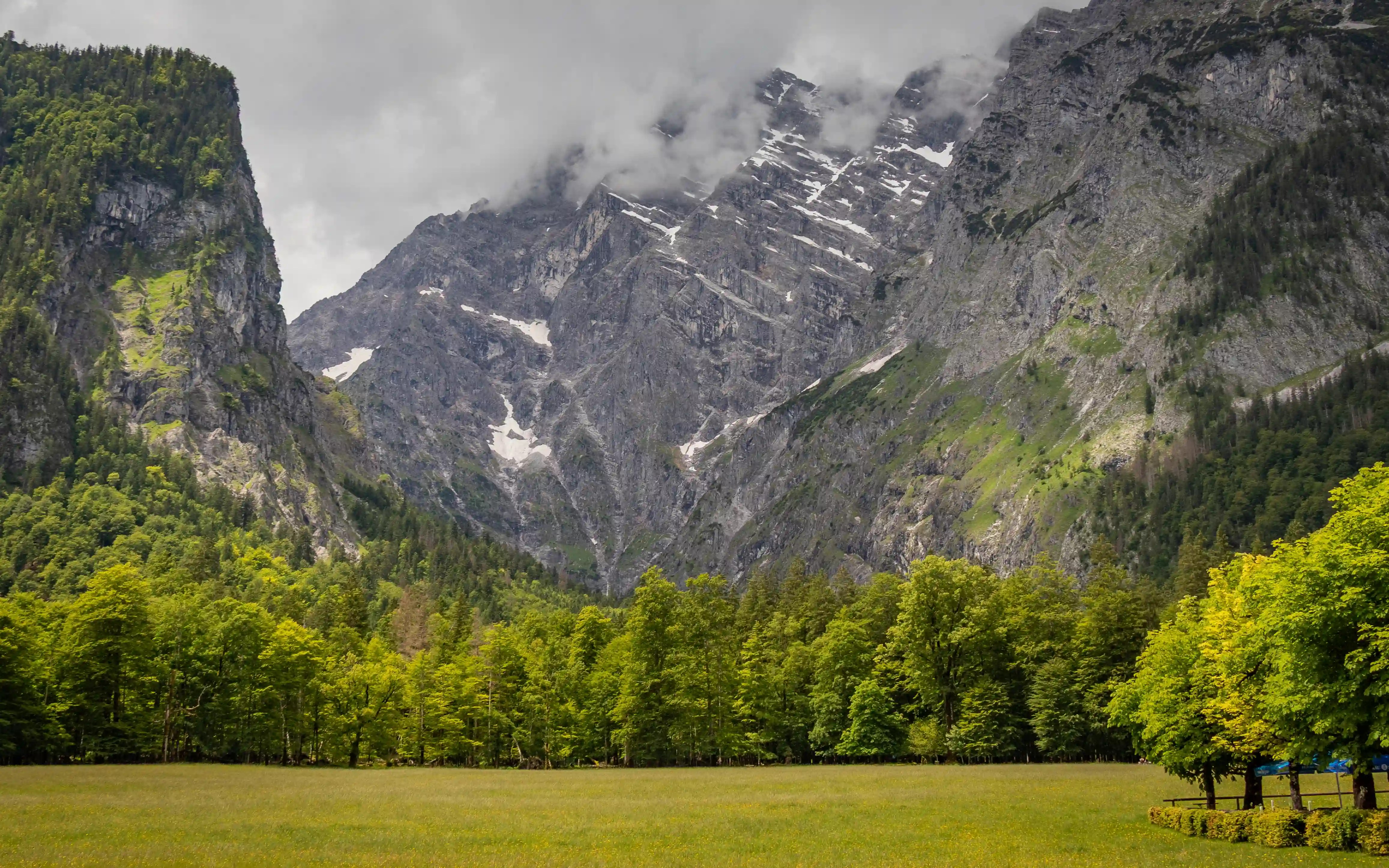 A wide alpine meadow stretches toward steep gray mountain peaks partially covered by clouds near St. Bartholomä.