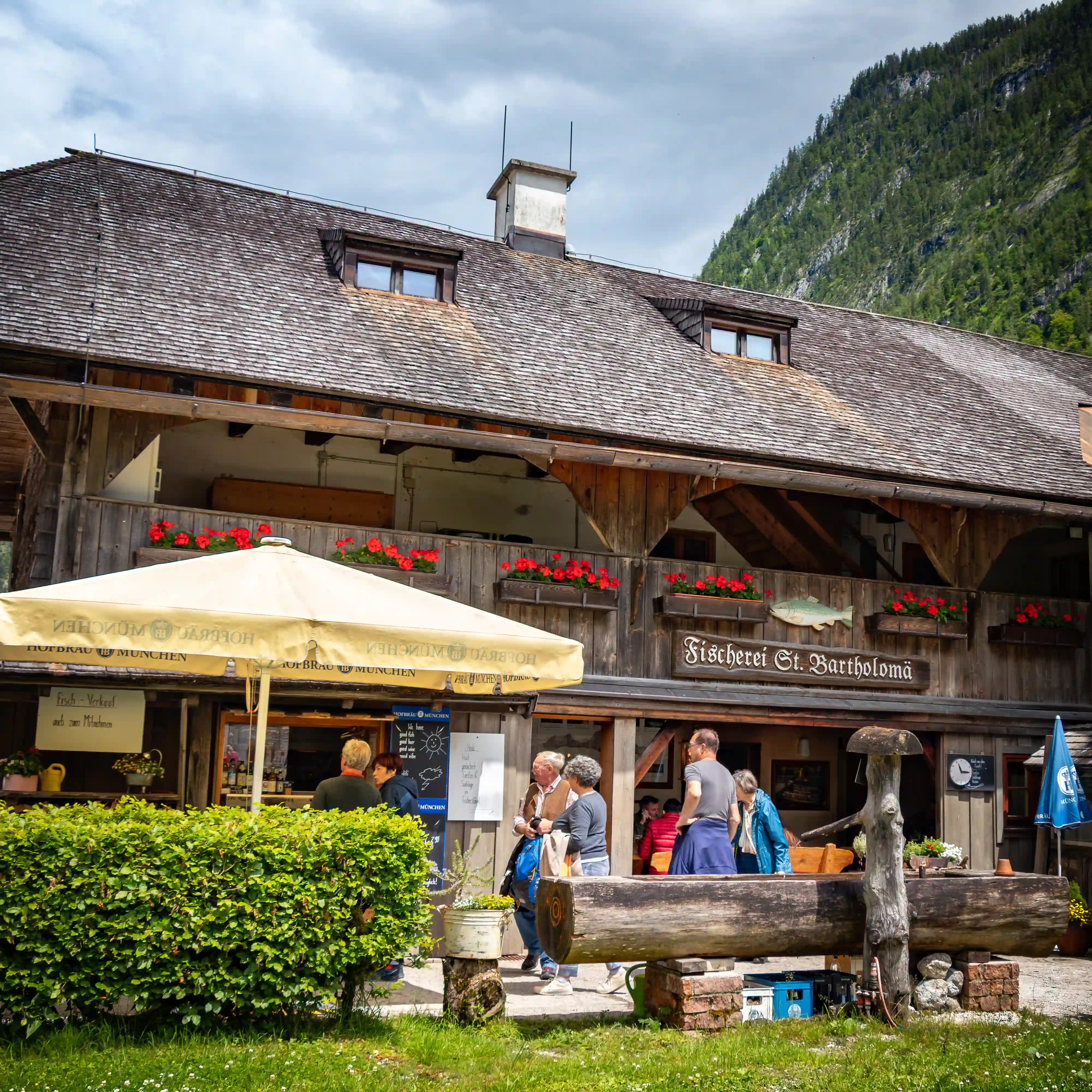 Visitors gather outside the Fischerei St. Bartholomä, a rustic wooden restaurant with flower boxes and outdoor seating.