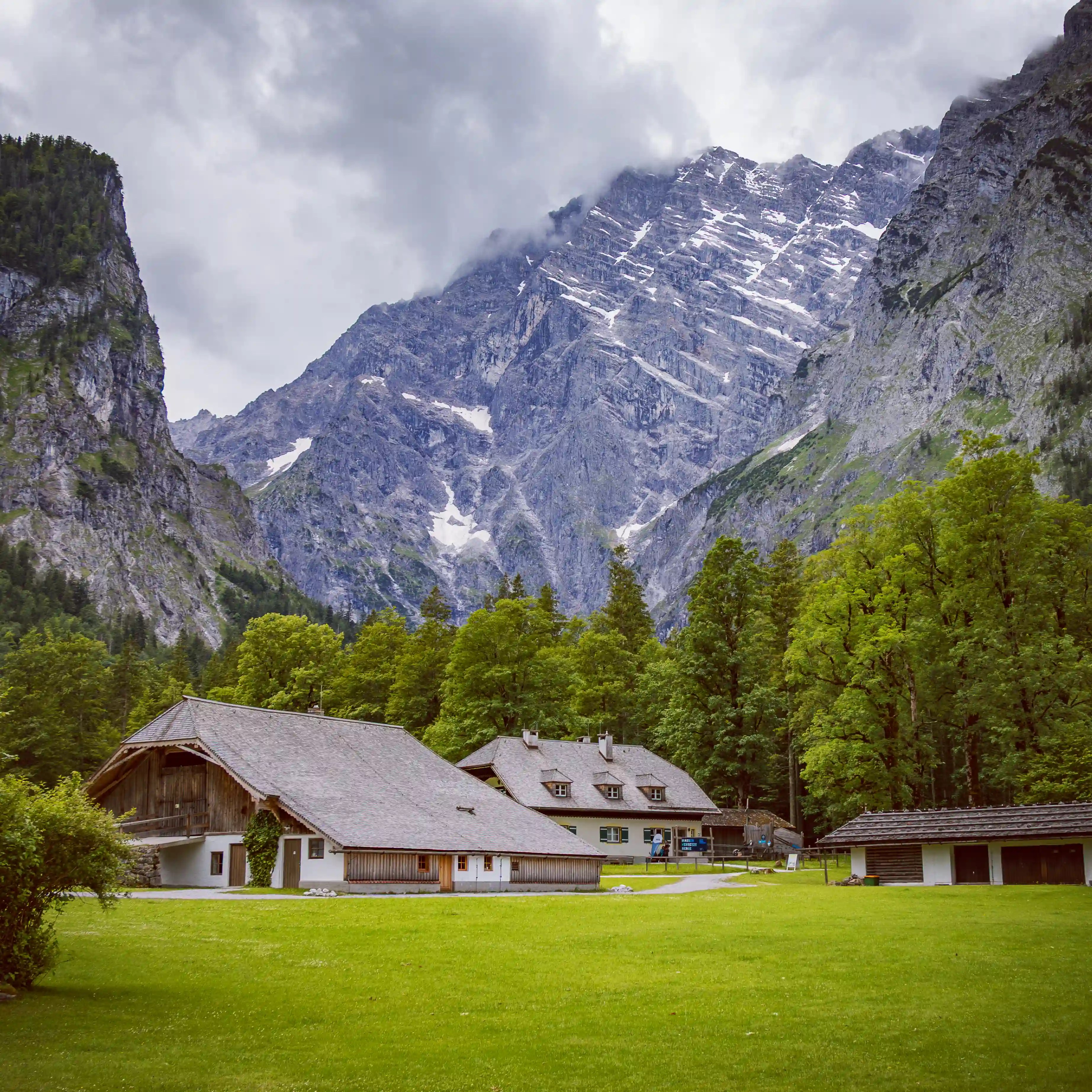 Low alpine buildings sit in a grassy clearing near St. Bartholomä with dramatic mountain cliffs rising behind them.
