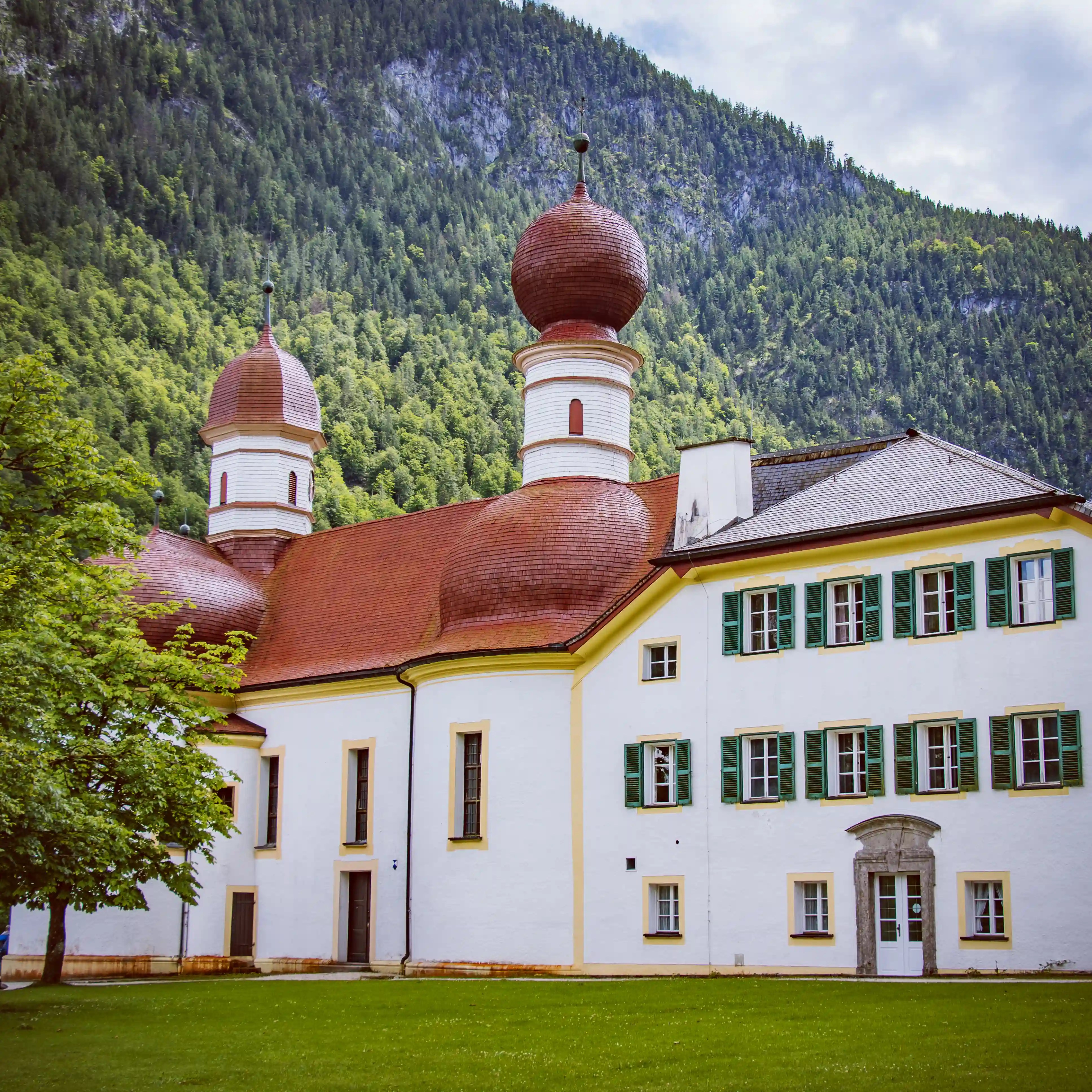 A close view of the white walls and red onion domes of the Church of St. Bartholomä against a forested mountainside.