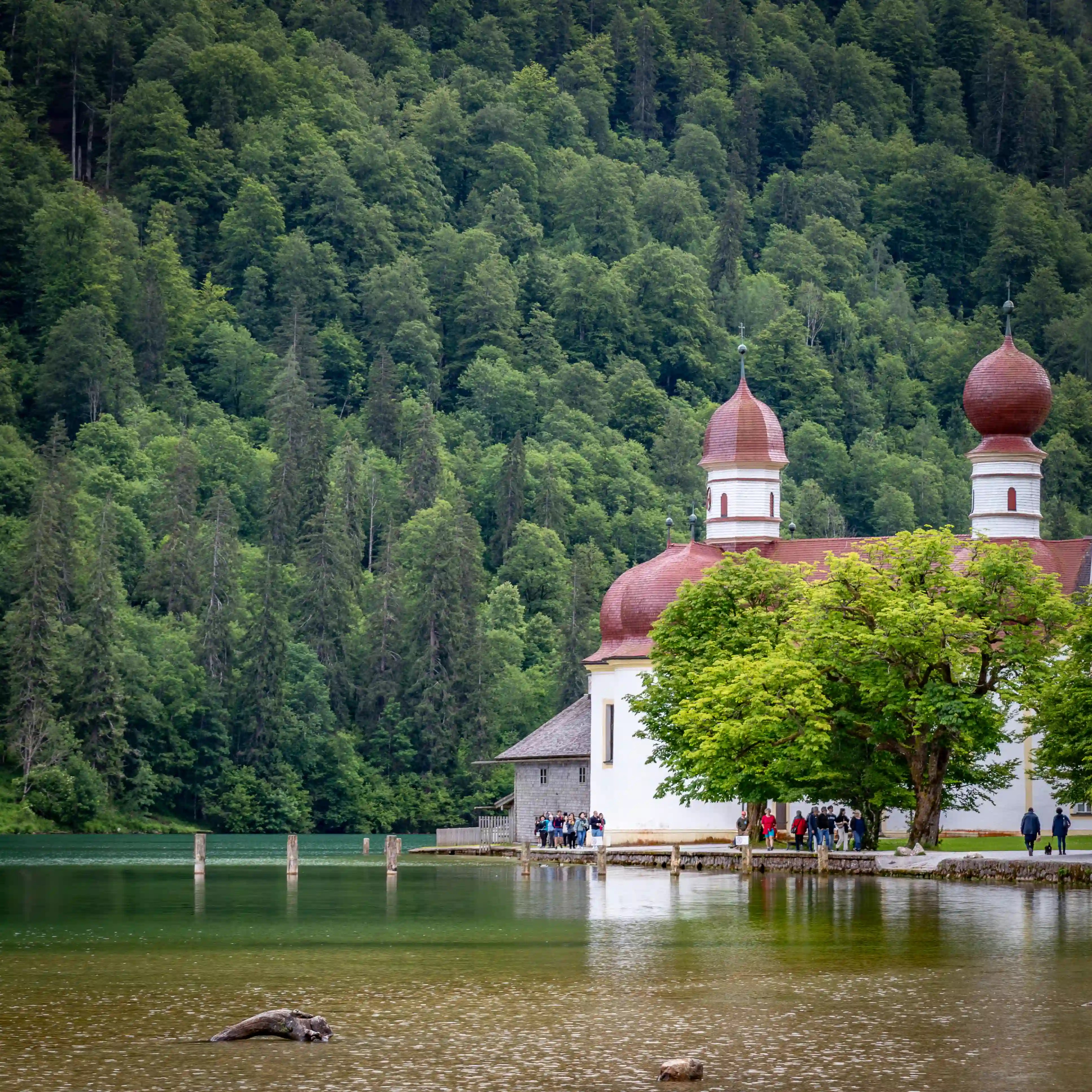 The Church of St. Bartholomä stands beside calm green water as visitors gather along the shoreline beneath dense forest.