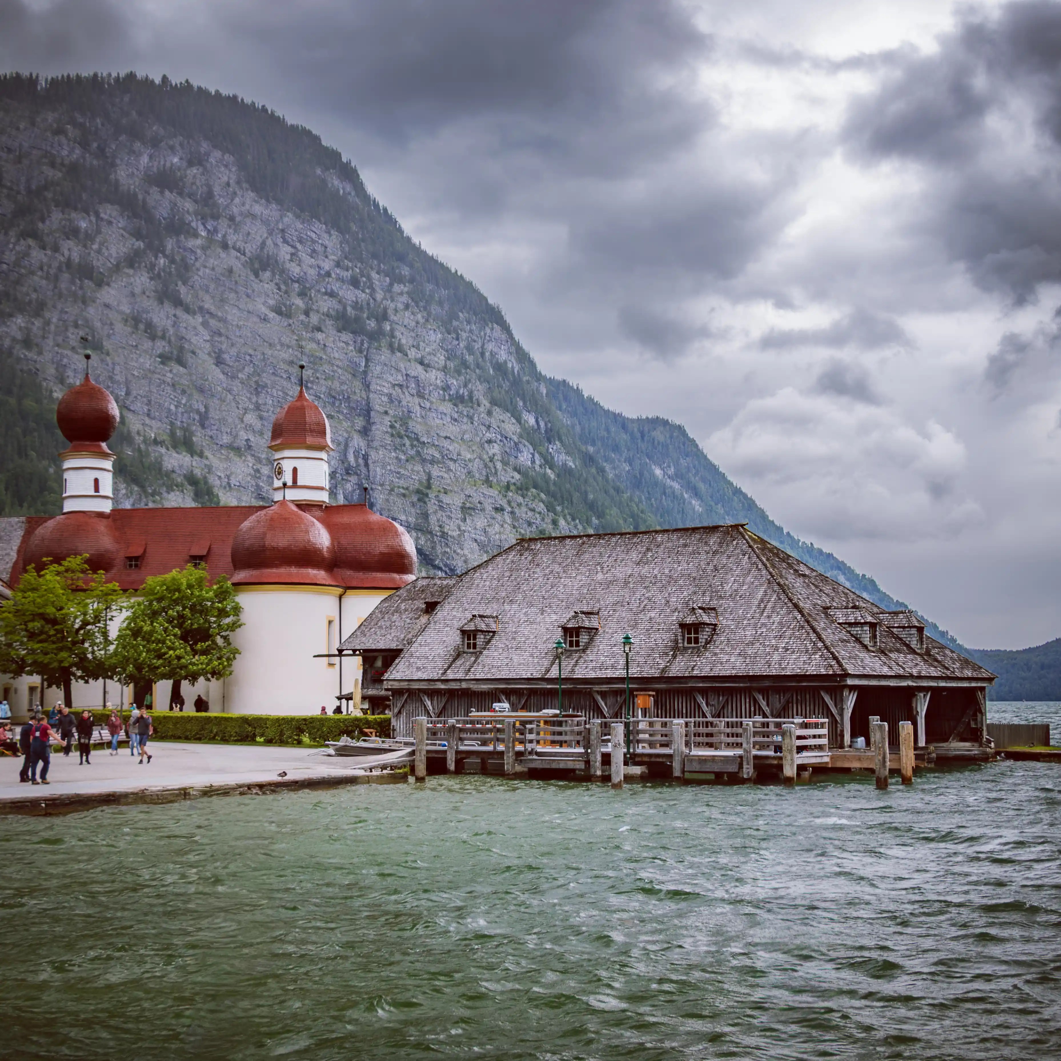 The red-domed pilgrimage church of St. Bartholomä stands beside a wooden boathouse on the edge of Königssee, with visitors gathered along the shore and mountains rising behind.