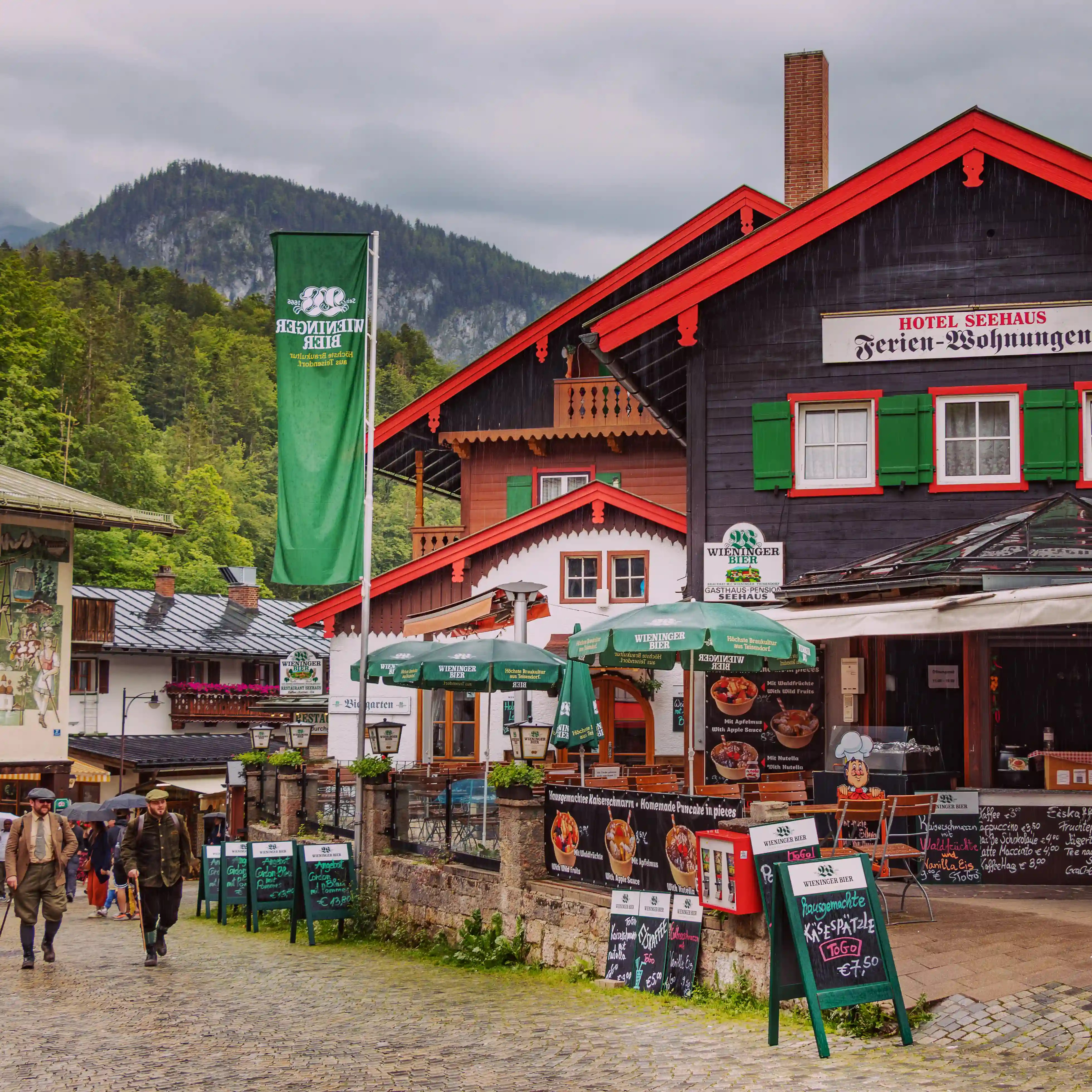 Colorful alpine buildings and restaurants line a cobblestone street near the Hotel Seehaus Königssee, with menu boards and pedestrians along the walkway.