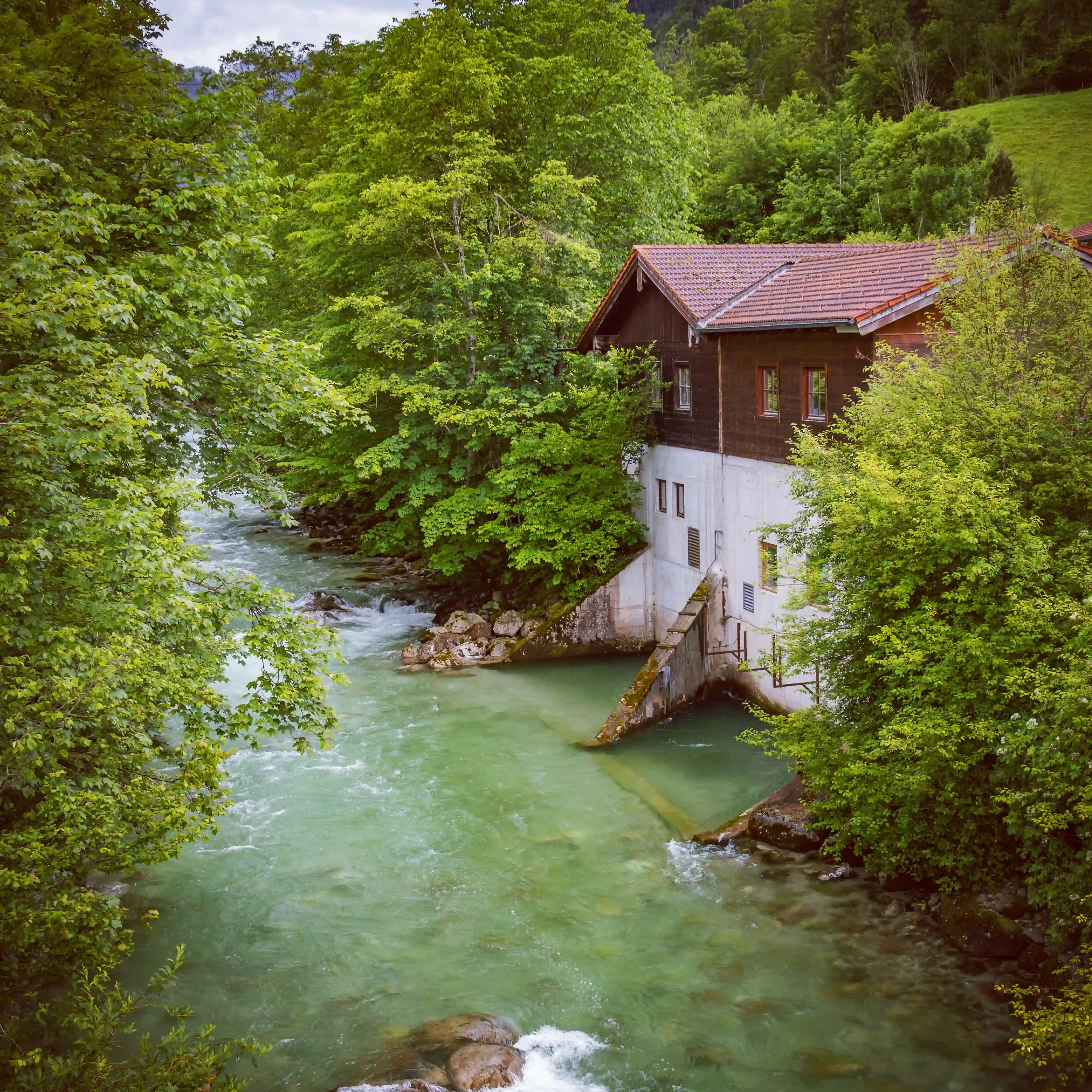 A historic wooden mill building sits beside a fast-flowing green mountain stream surrounded by dense forest near Königssee.