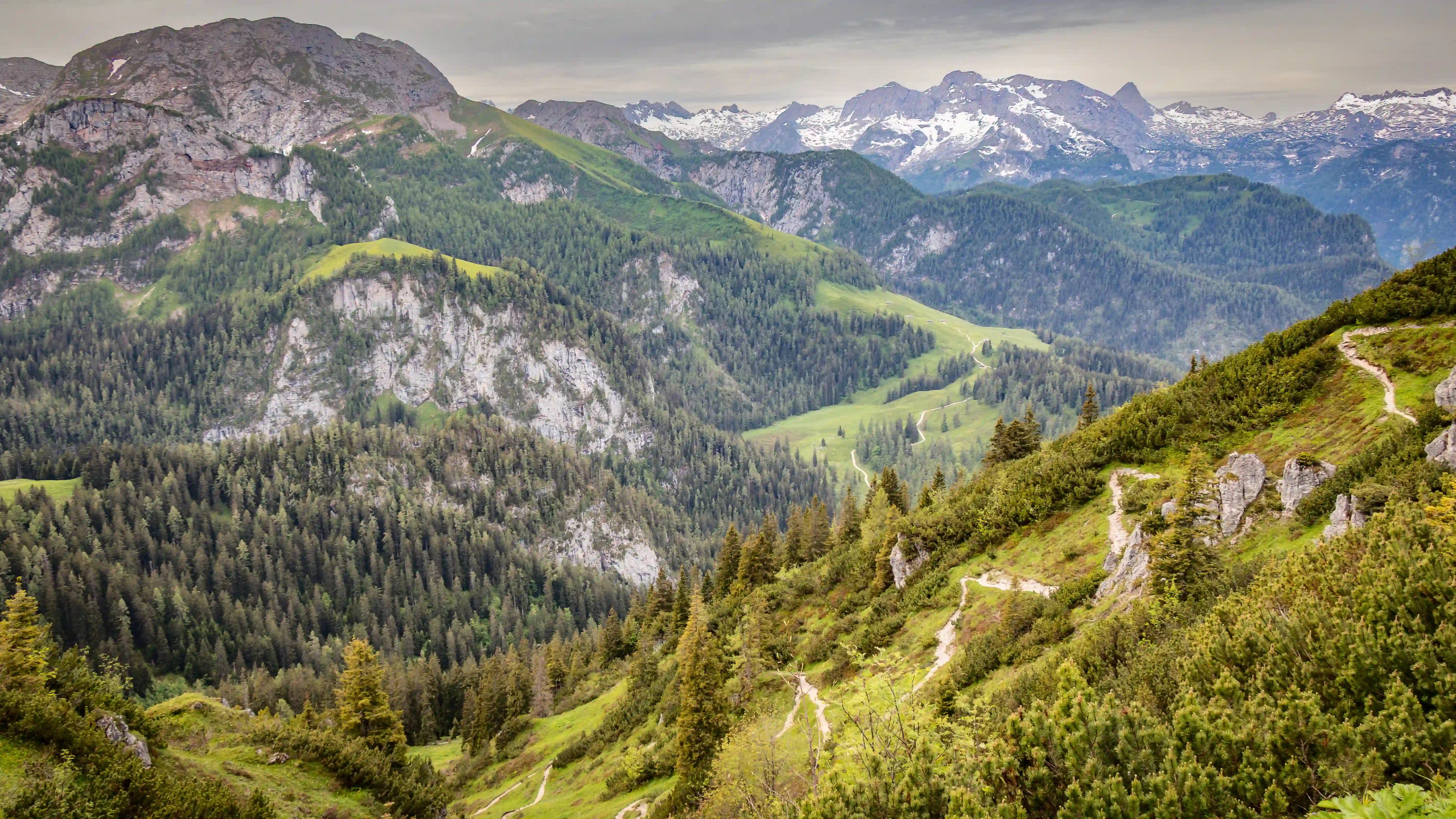 Steep rocky cliffs and forested ridges descend into a wide alpine valley, with snow-covered mountains rising in the distance.