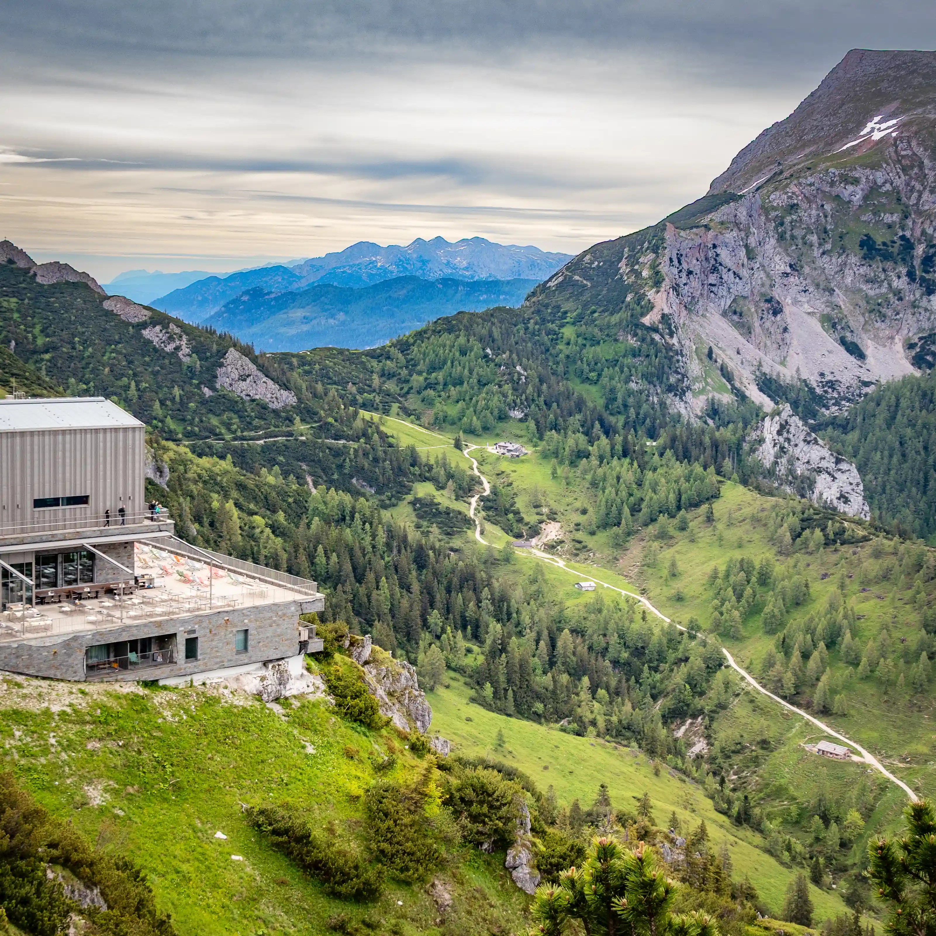 A modern mountain building with a terrace clings to a steep grassy slope, overlooking a vast alpine valley crossed by narrow hiking trails.