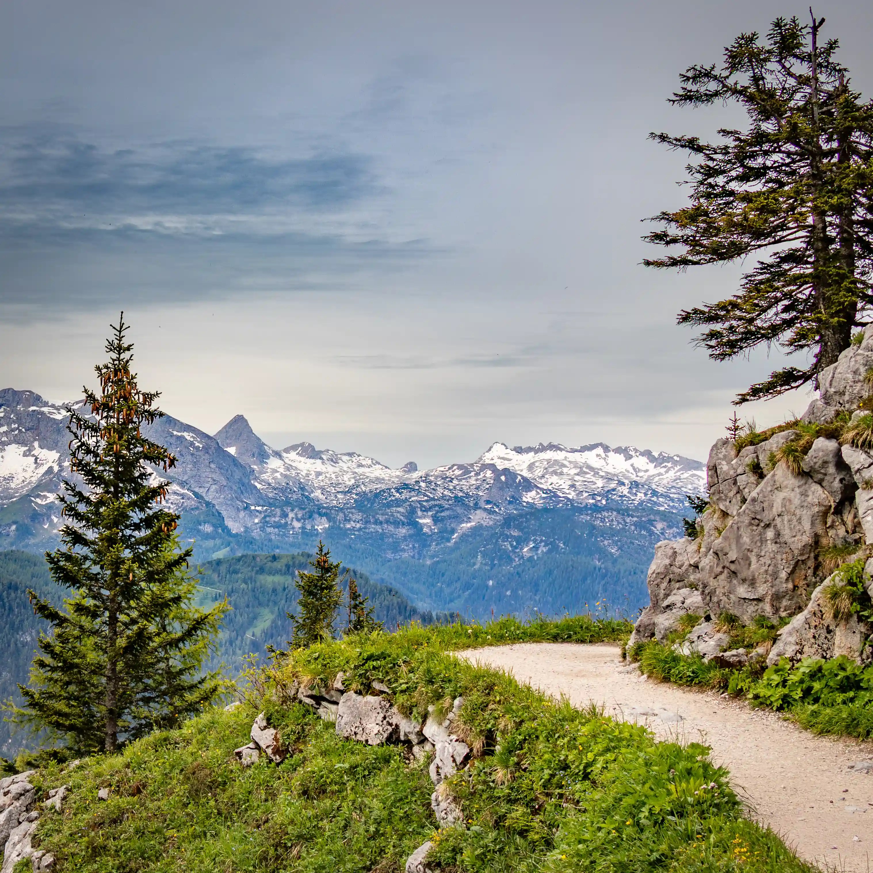 A narrow gravel path curves along a rocky hillside bordered by grass and small trees, with distant mountain peaks visible beyond.