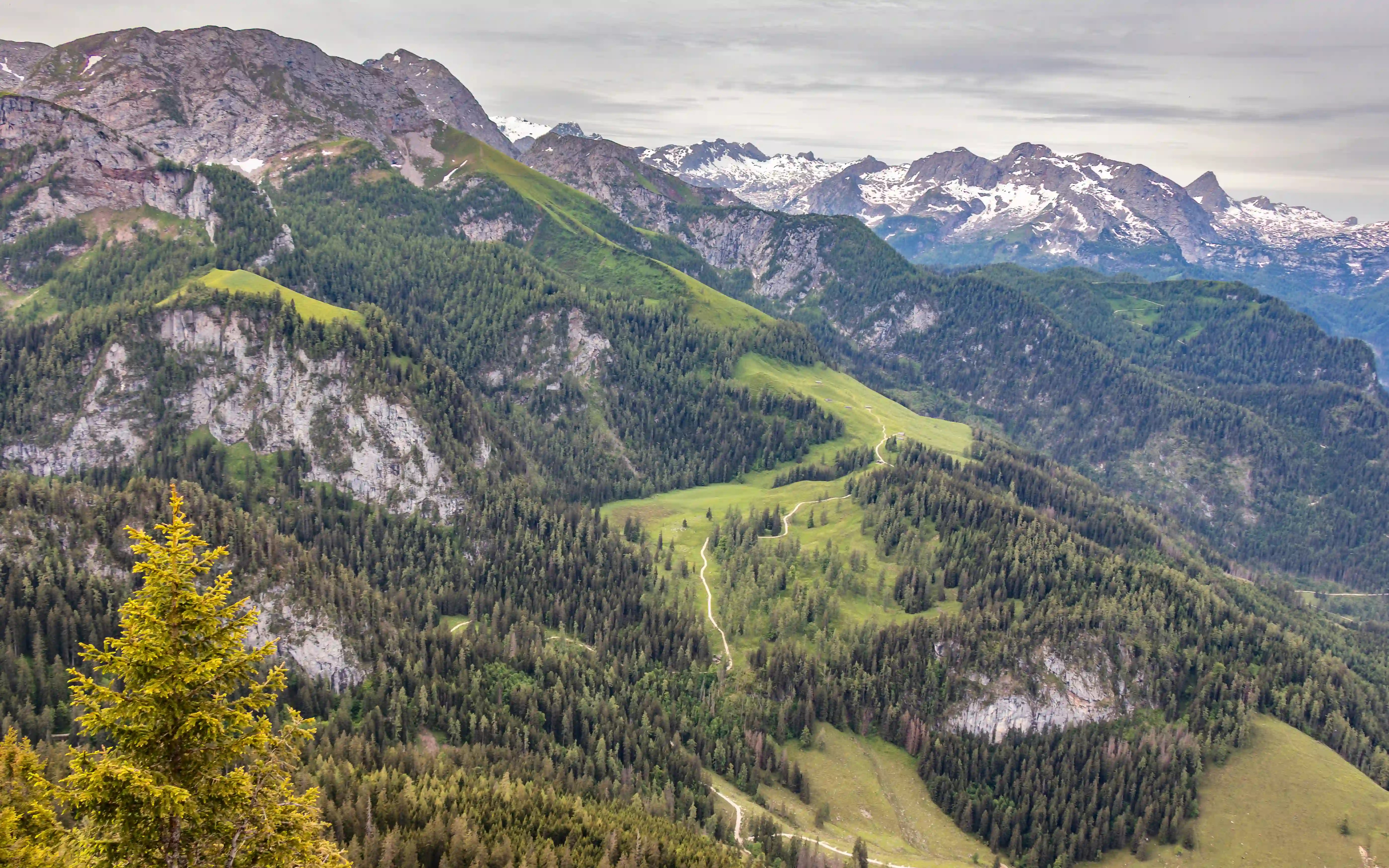 Green alpine ridges and forested slopes roll across the landscape, with pale hiking paths tracing their way through the terrain.