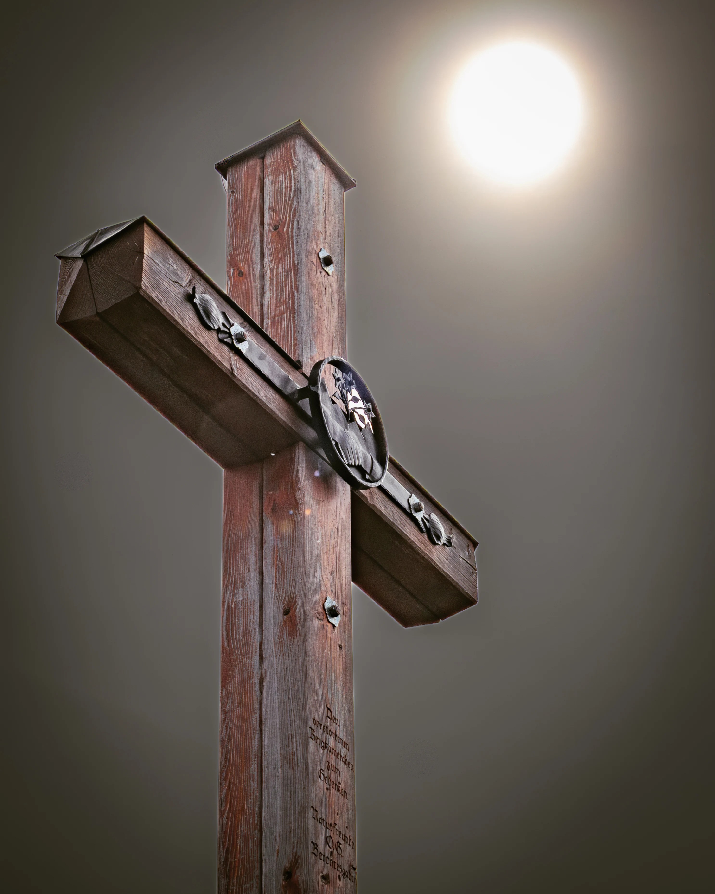 A wooden summit cross stands against a muted sky with the sun glowing behind it.