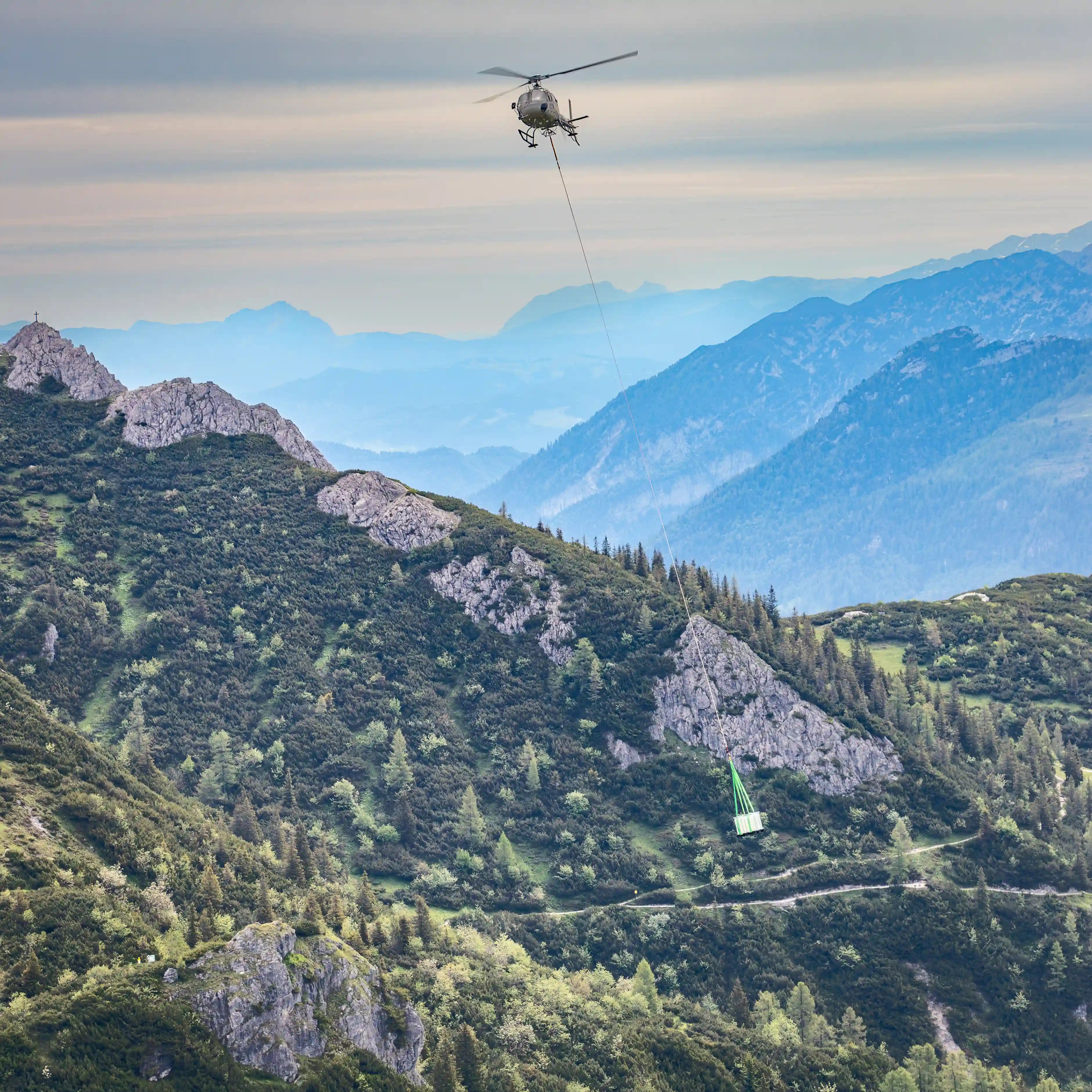 A helicopter carries a suspended load over forested mountain slopes above the valley.
