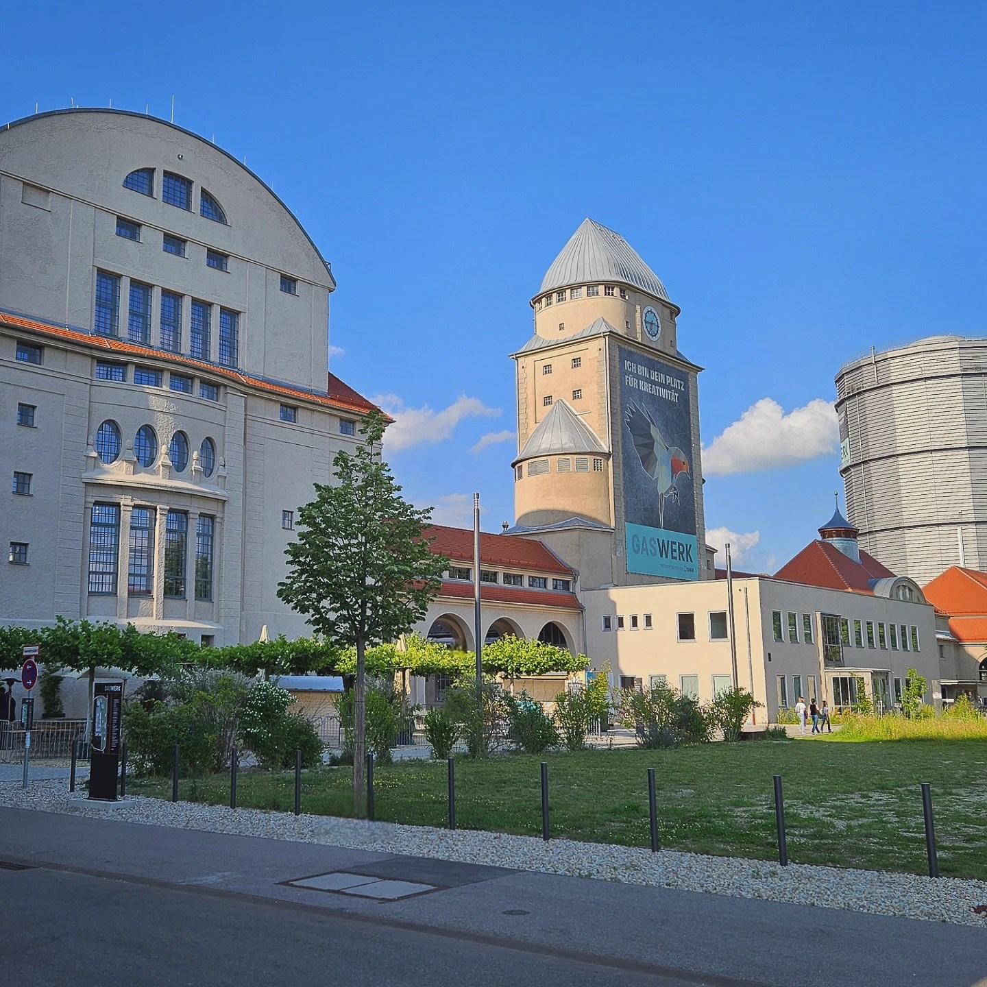 The Gaswerk Augsburg complex with historic industrial buildings, towers, and open green space under a bright blue sky.