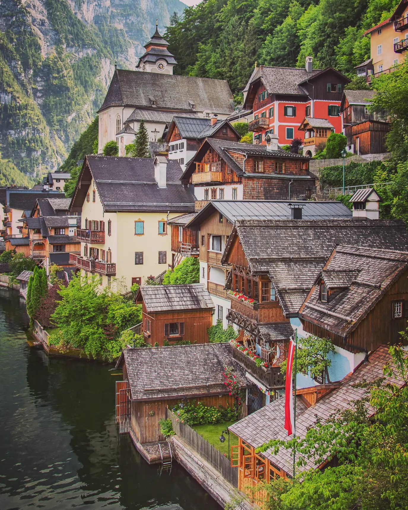 Colorful wooden houses climbing a hillside in Hallstatt with a church and forested cliffs behind them.