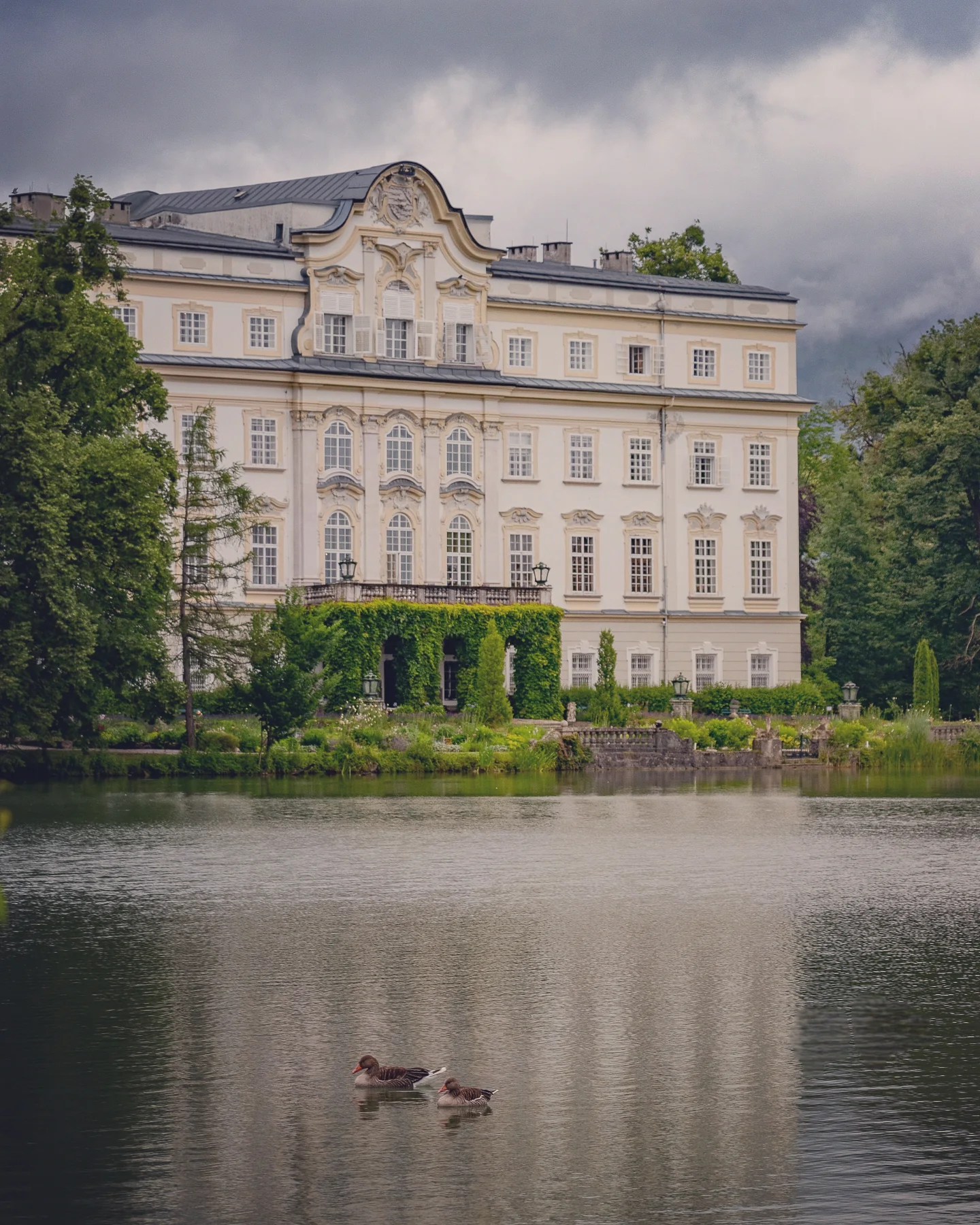 A pale yellow and white palace with ornate windows sits across a calm lake, reflecting in the water.