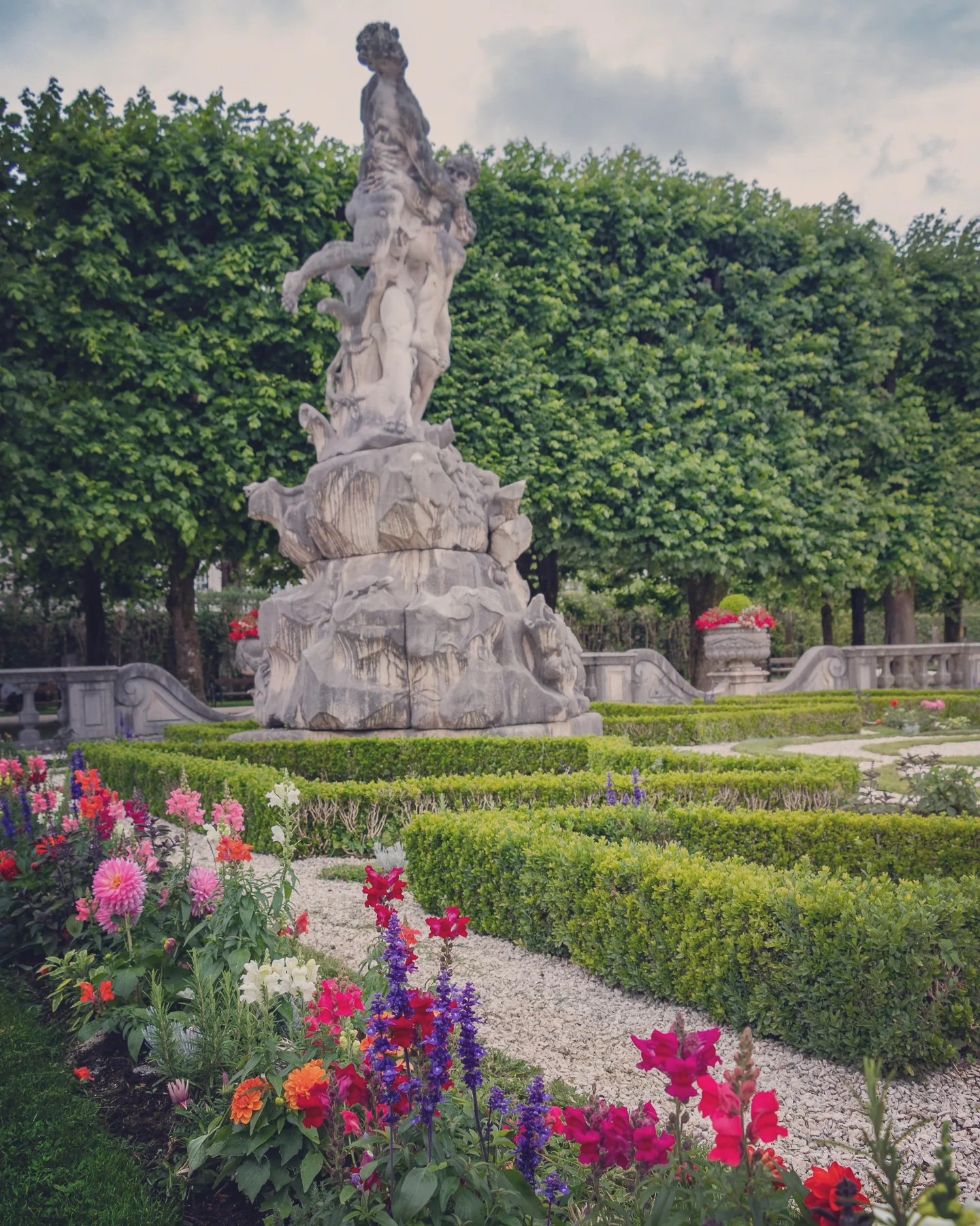 A stone statue rising from a rocky base in Mirabell Gardens, surrounded by colorful flower beds and neatly trimmed hedges.