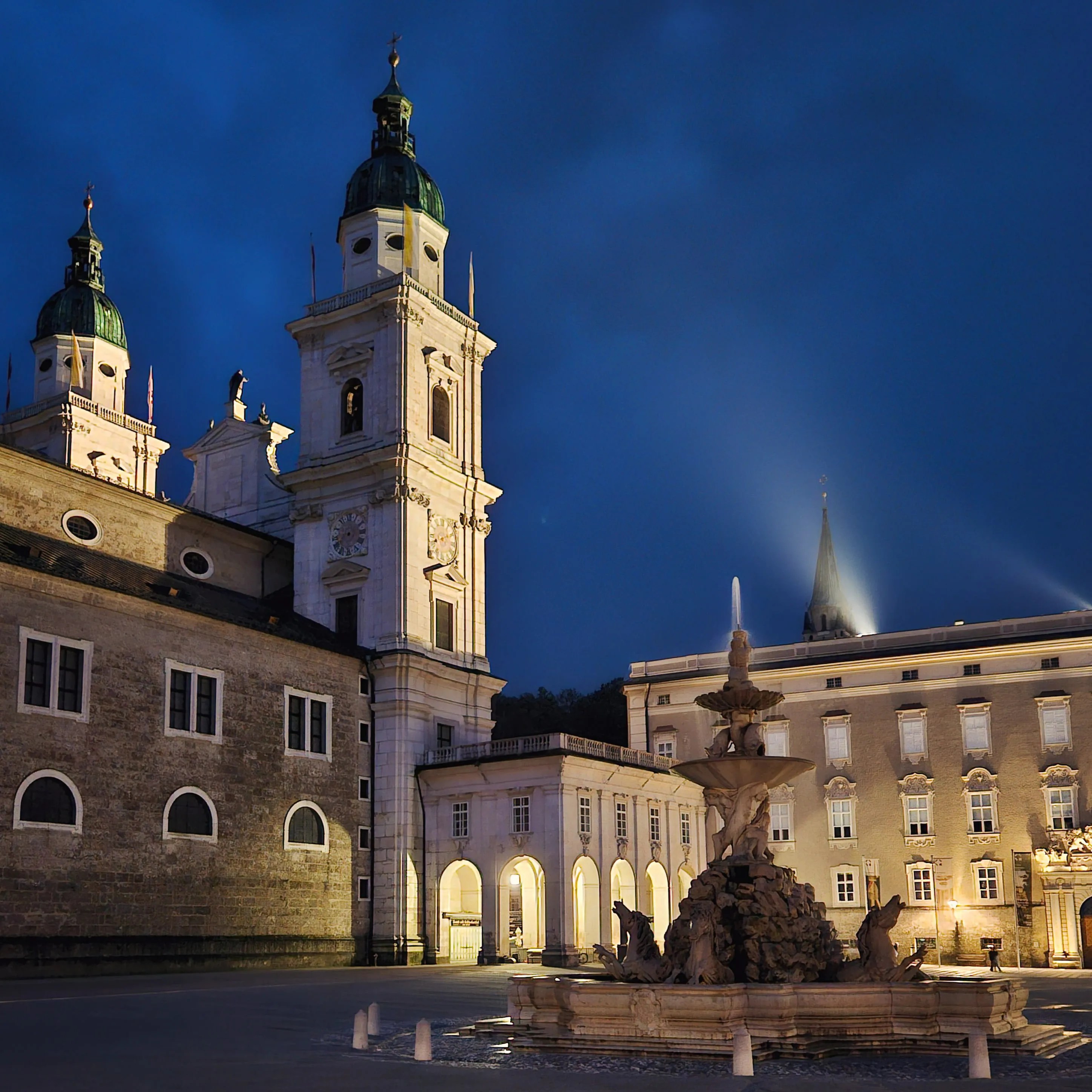 Salzburg Cathedral and the Residenz Fountain are illuminated at night, with the Baroque buildings glowing against a deep blue sky.