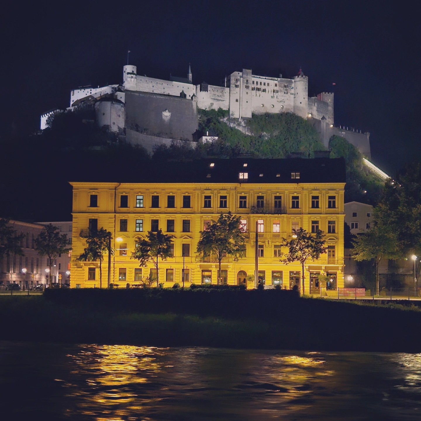 Hohensalzburg Fortress sits illuminated on a hill at night above the Salzach River, with a glowing yellow building reflected in the water below.