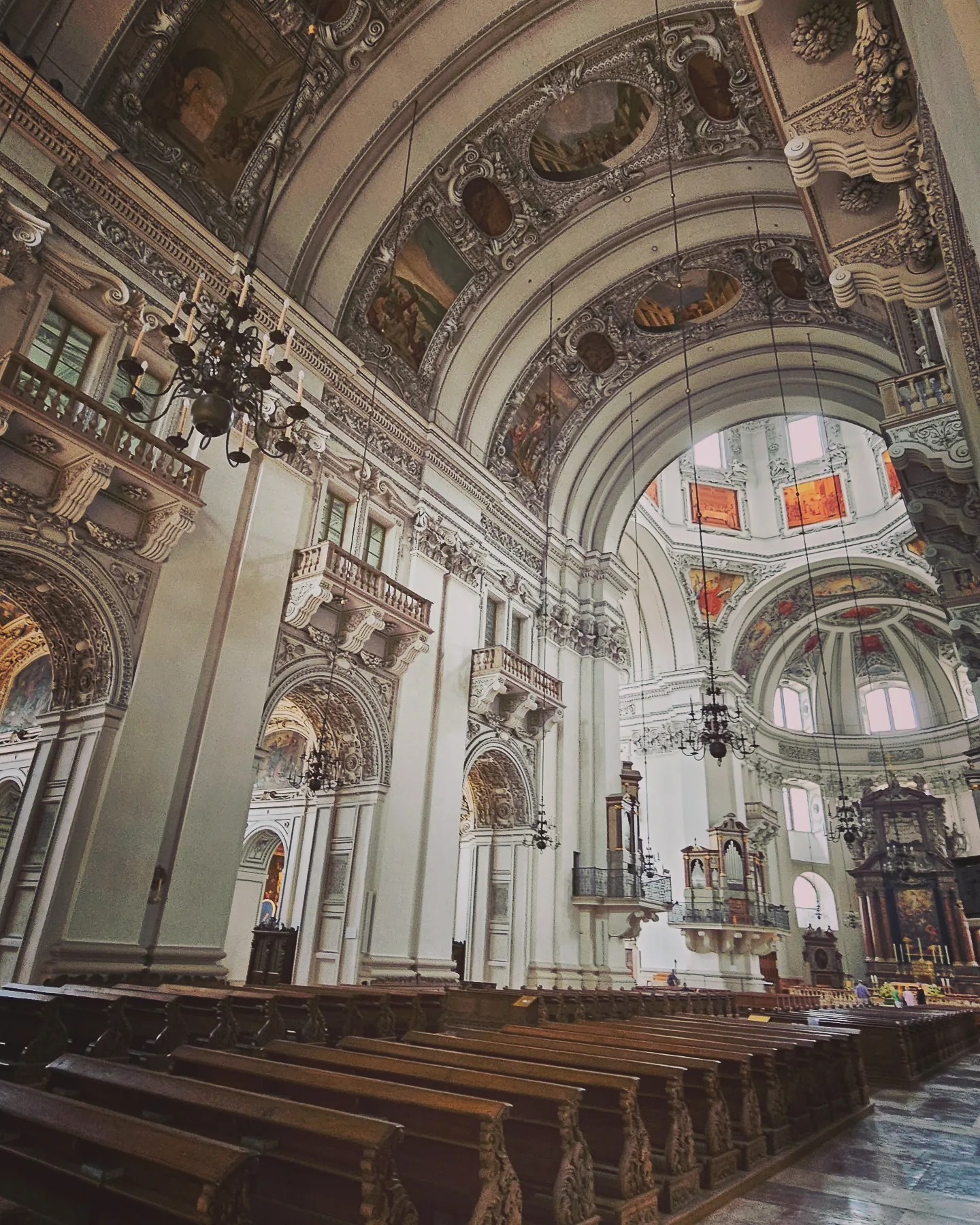 Rows of wooden pews line a side aisle inside Salzburg Cathedral, with arched galleries, chandeliers, and frescoed ceilings above.