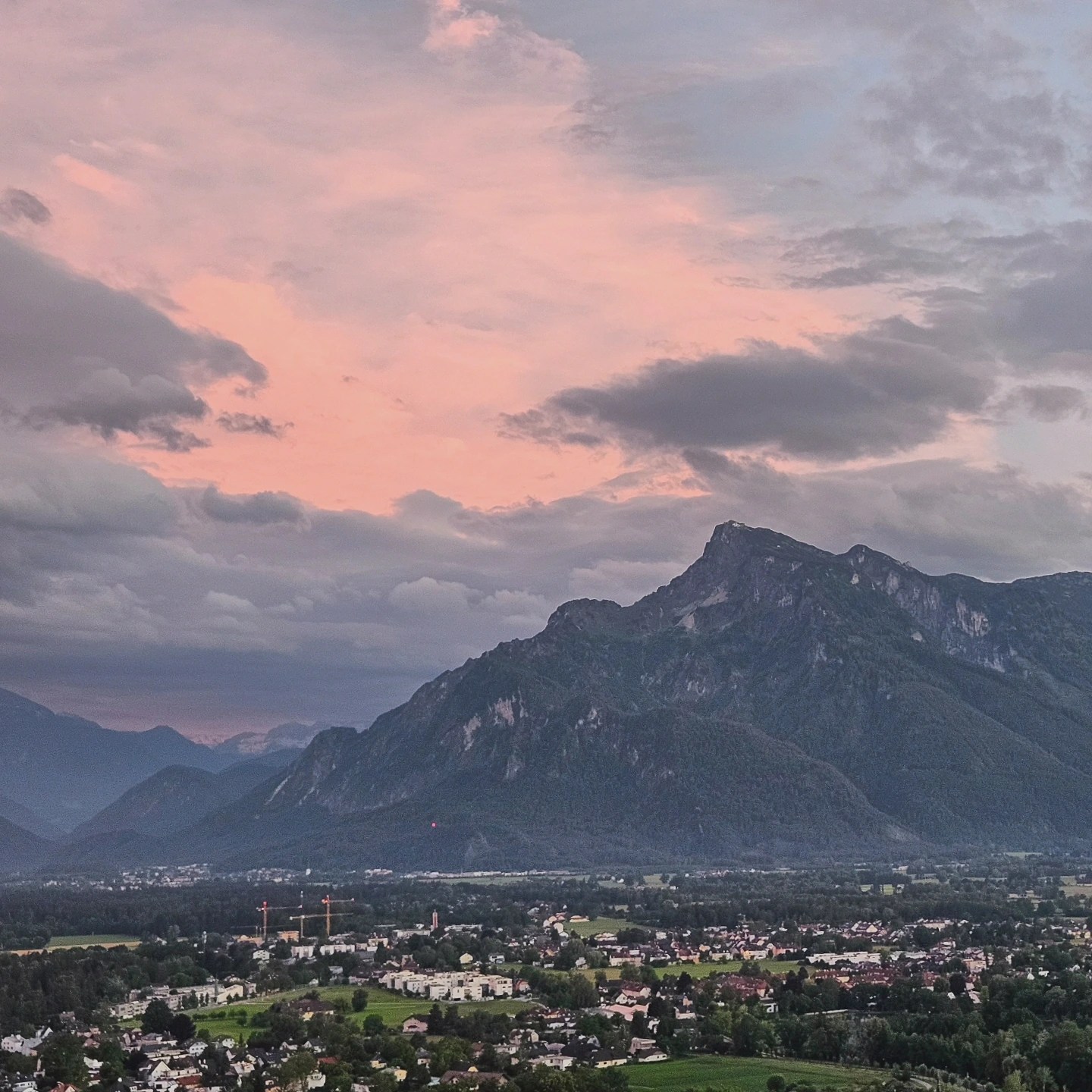 A broad valley view at sunset with layered mountains, scattered towns, and a dramatic pink and gray sky seen from Hohensalzburg Fortress.