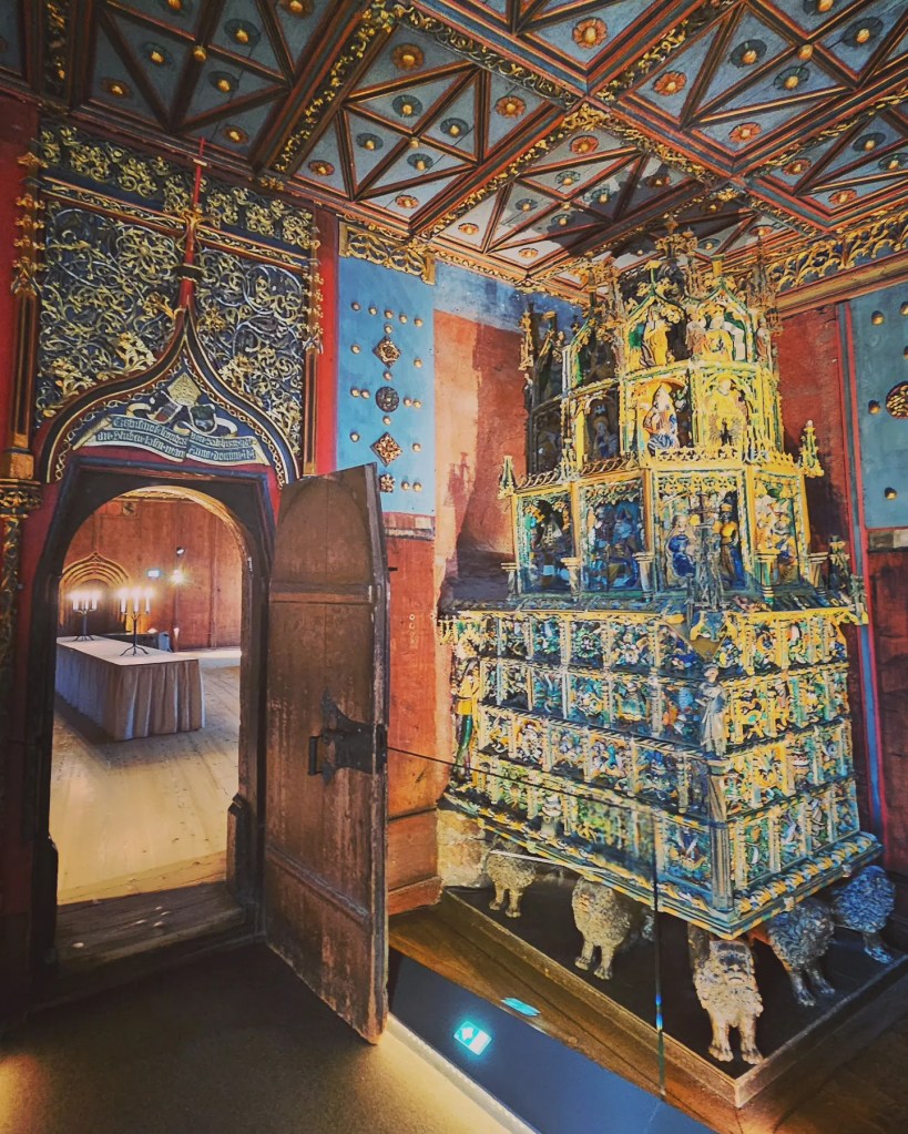 An ornate gilded medieval stove displayed in a richly decorated room inside Hohensalzburg Fortress, with painted walls and a carved wooden doorway.