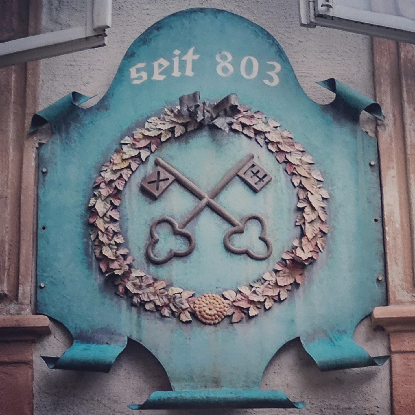 A turquoise crest with crossed keys and the text “seit 803” is mounted above an arched entrance at St. Peter’s Stiftskulinarium.