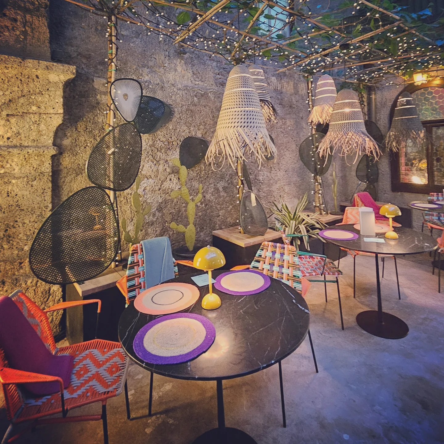 Small round tables with colorful chairs, woven placemats, and hanging lanterns are arranged in a stone-walled courtyard dining area.