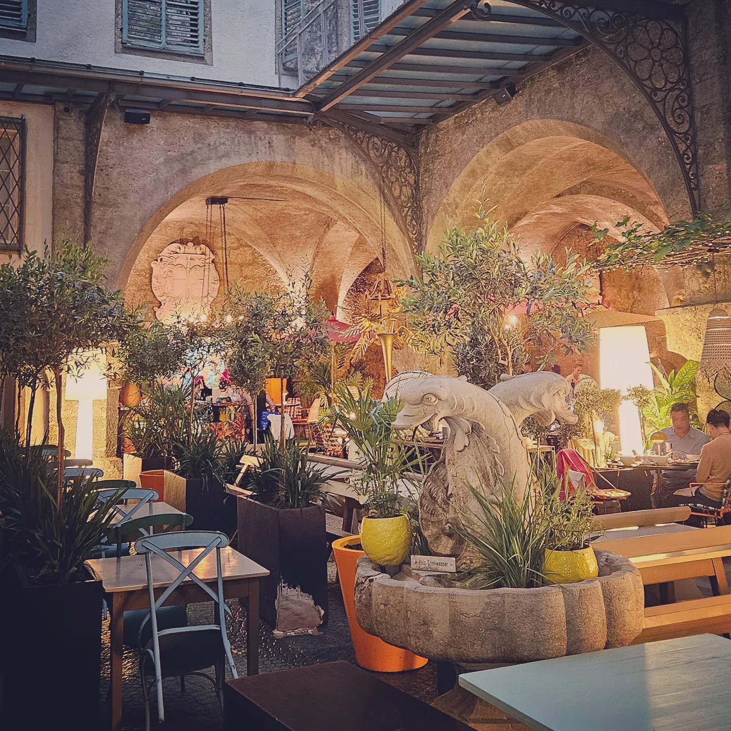 A stone fountain surrounded by plants sits beneath arched ceilings in the softly lit courtyard of St. Peter’s Stiftskulinarium.