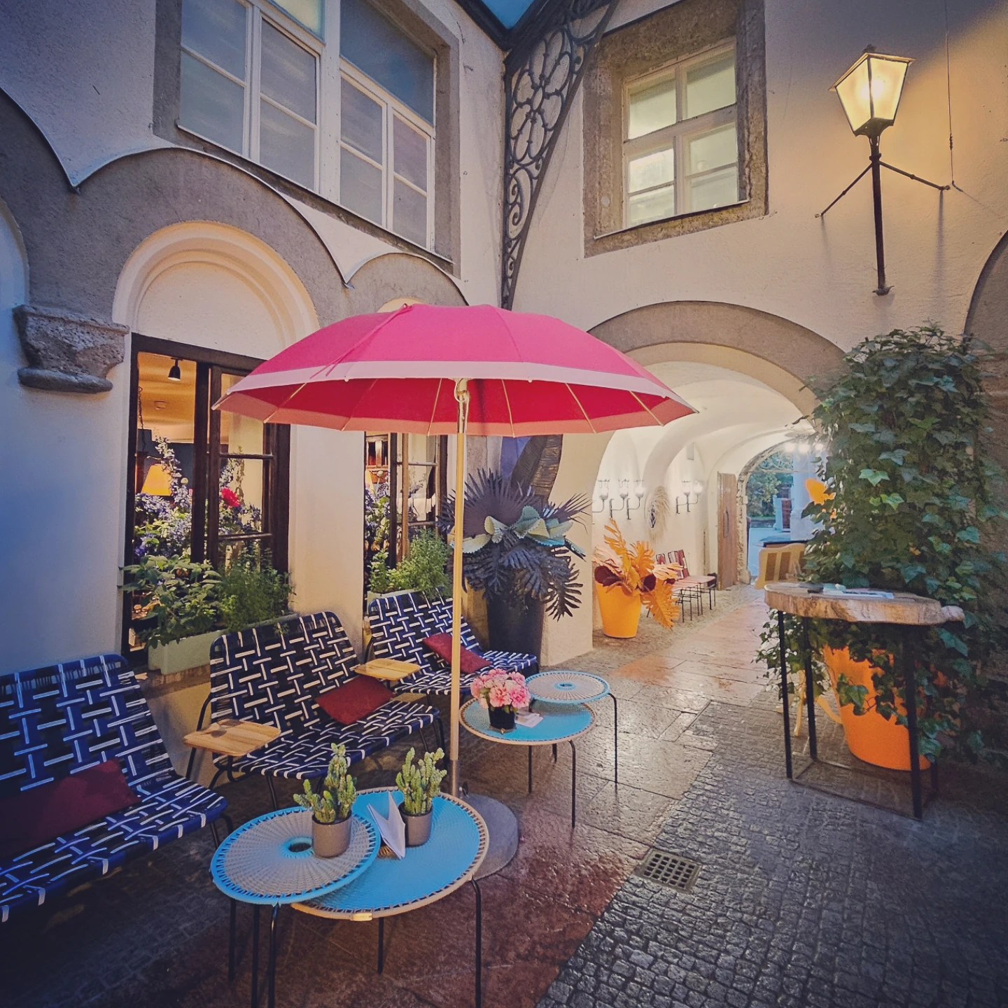 Outdoor seating with blue patterned chairs, small round tables, and a pink umbrella beneath arched walls in the courtyard of St. Peter’s Stiftskulinarium in Salzburg.