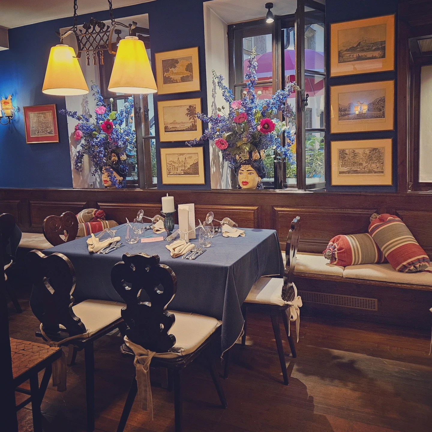 An elegant dining room at St. Peter’s Stiftskulinarium with a set table, wooden chairs, framed artwork, and large floral arrangements near a window.