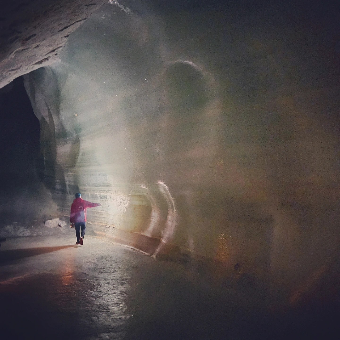 A guide walks along a smooth ice wall inside Eisriesenwelt, lightly touching the frozen surface as light reflects across it.