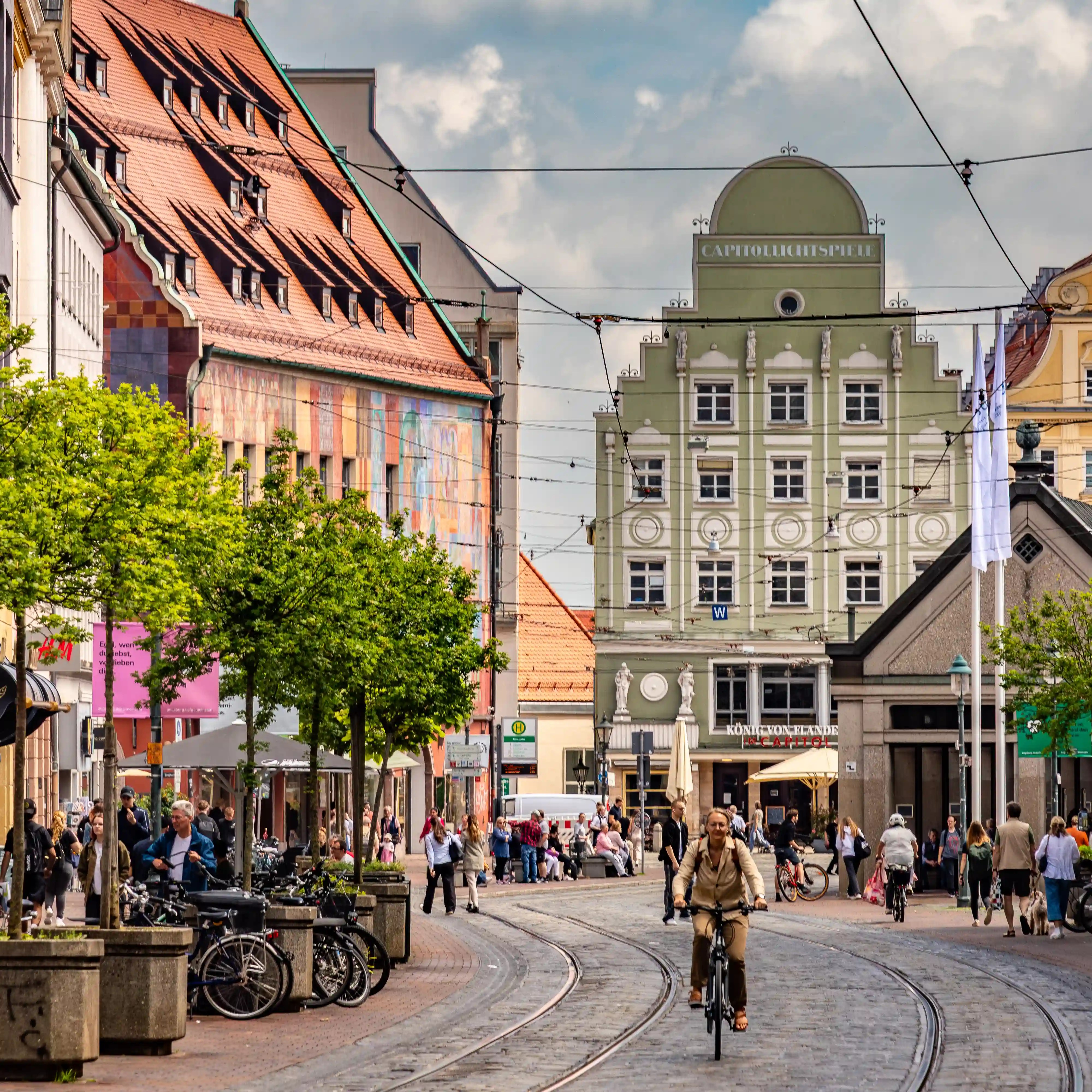 A cyclist rides along tram tracks through a busy street in Augsburg lined with historic buildings and cafés.